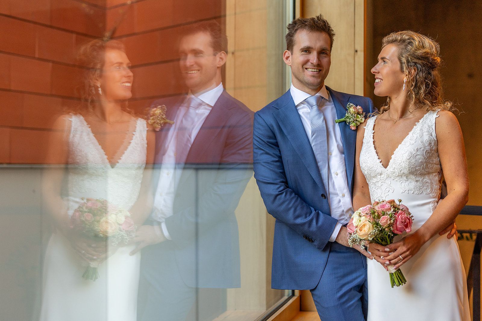 Bruid en bruidegom lachend, poserend samen op hun trouwdag. Zij houdt bloemen vast, hij draagt een blauw pak.