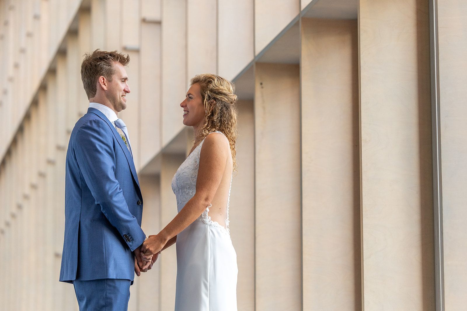 Een stel houdt elkaars hand vast en lacht naar elkaar. Man in blauw pak, vrouw in witte jurk. Interieur met raam.