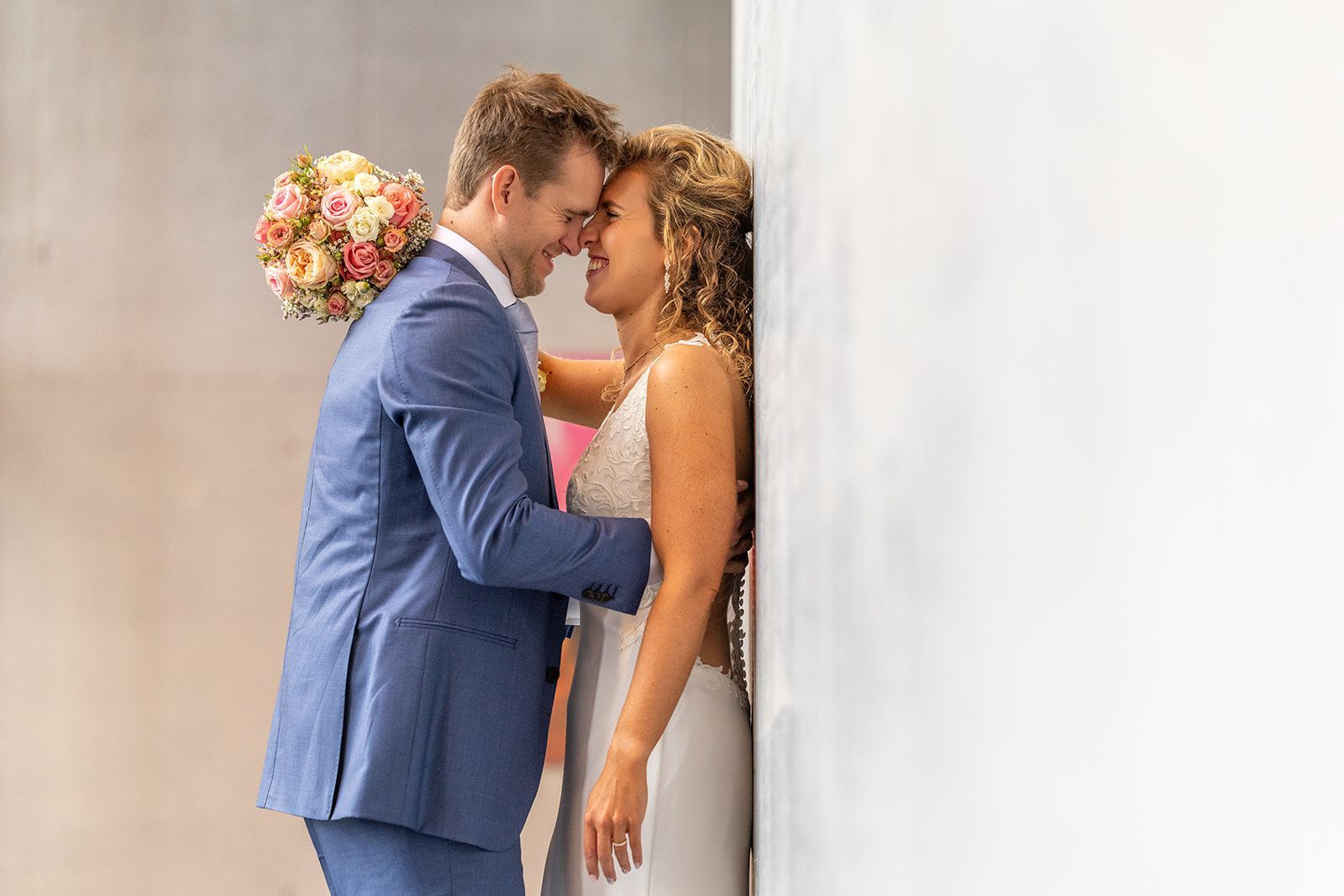 Het bruidspaar omhelst elkaar, de bruid leunt tegen een muur met een boeket in haar handen. De bruidegom draagt een blauw pak.
