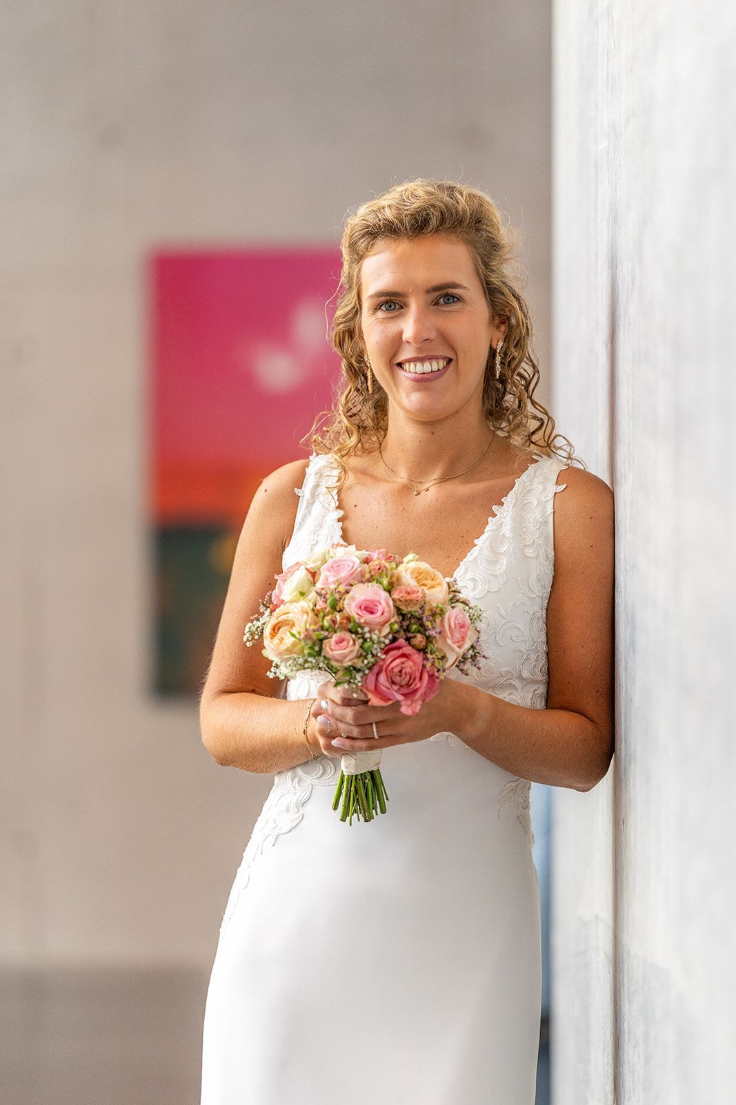 Bruid in witte jurk met boeket, leunend tegen een witte muur. Lachend. Roze en oranje kunstwerk op de achtergrond.