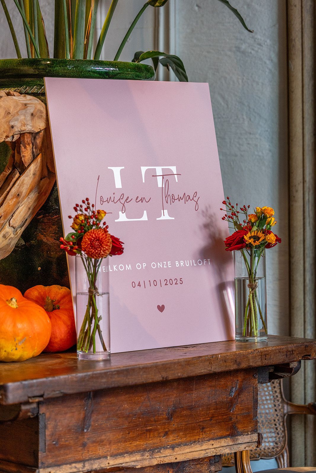 Roze bord met bloemendecoratie, pompoenen en een houten tafel, met de tekst