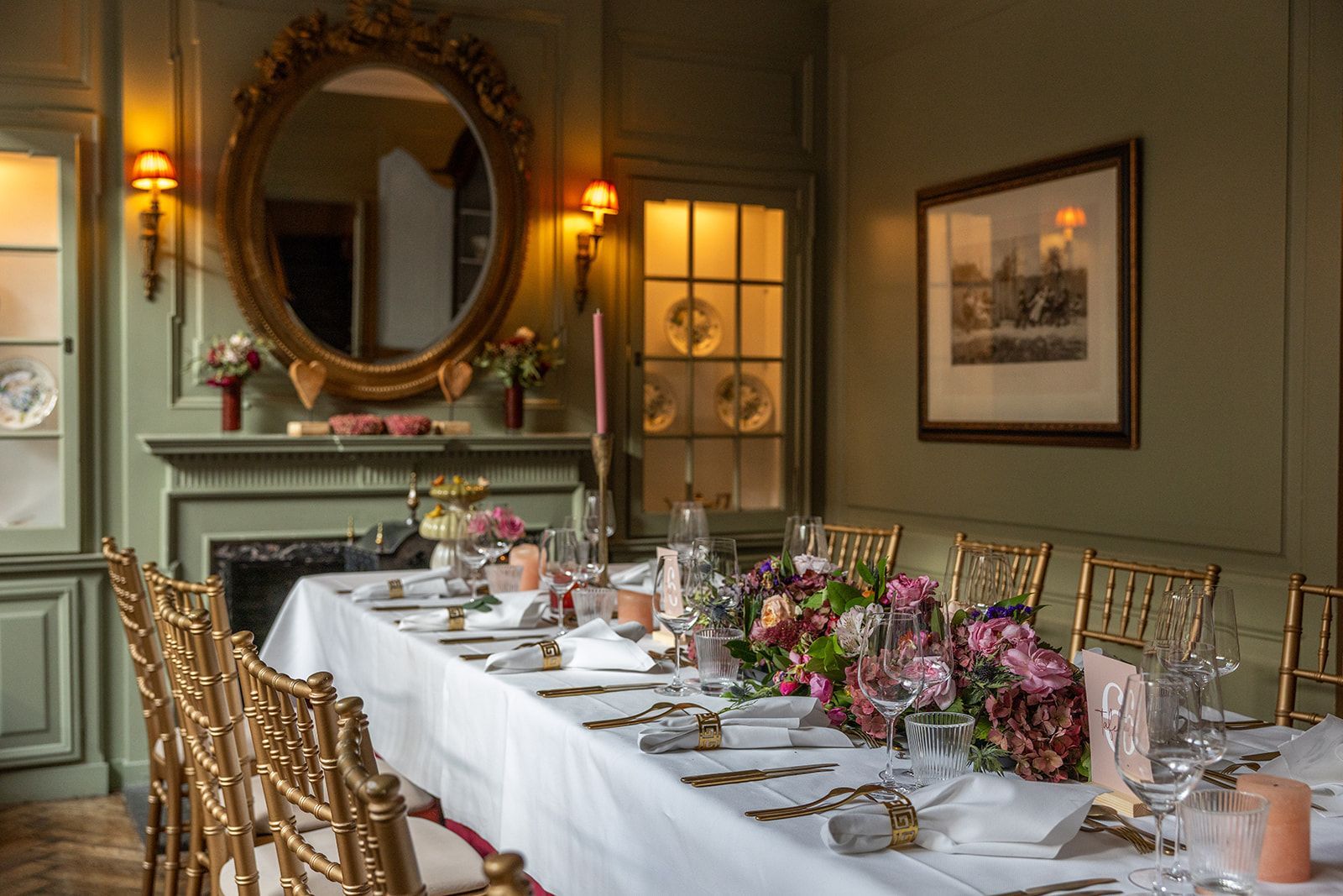 Elegante eetkamerset voor een maaltijd. Lange tafel, gouden stoelen, een bloemstuk, een ovale spiegel en kunstwerken. Groene muren.