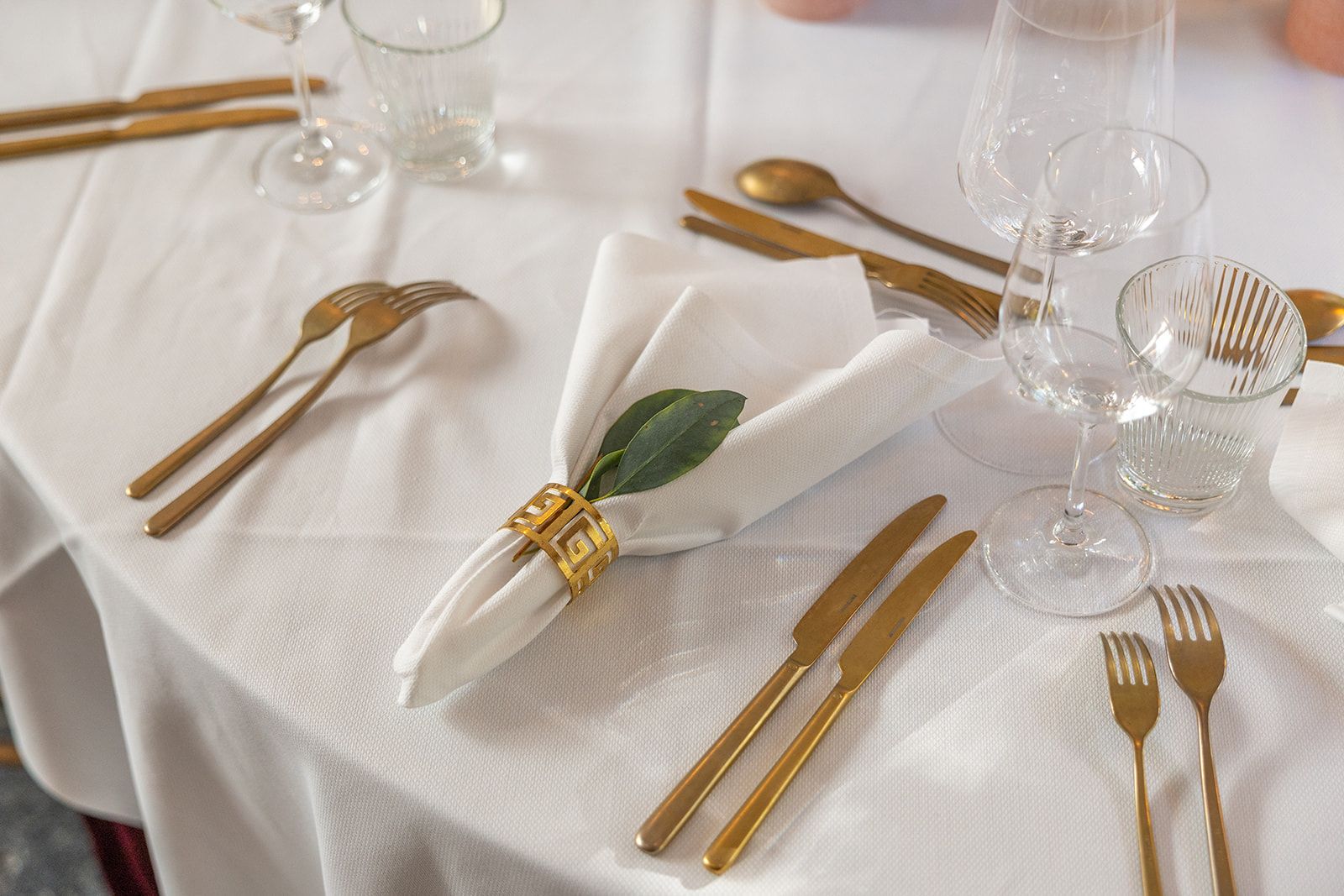 Tafeldekking met gouden zilverwerk, witte servetten en waterglazen op een wit tafelkleed.