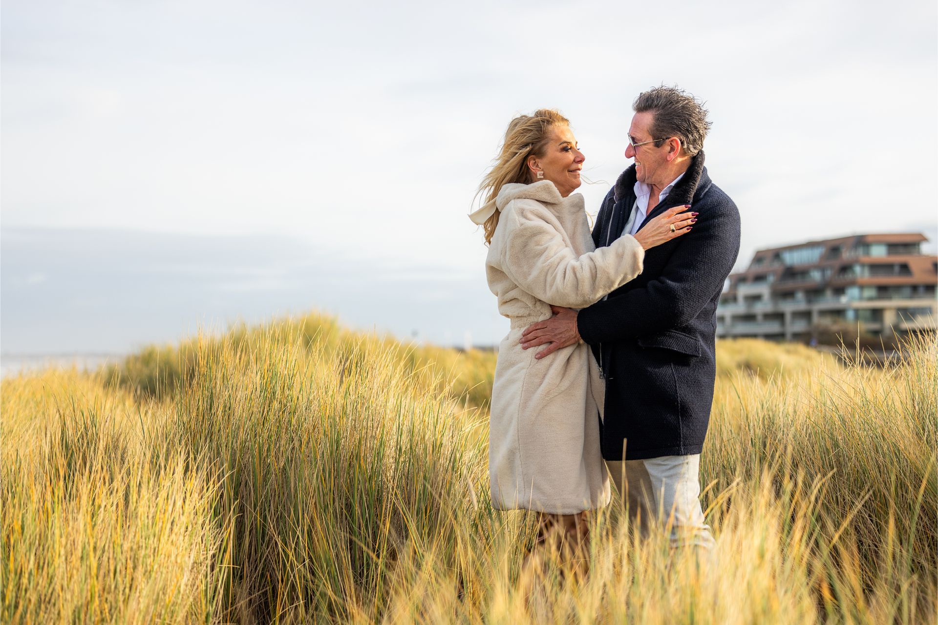 Een paartje omhelst elkaar in het hoge gras, glimlachend. Het strand en het gebouw op de achtergrond. Zonnig, bewolkt.