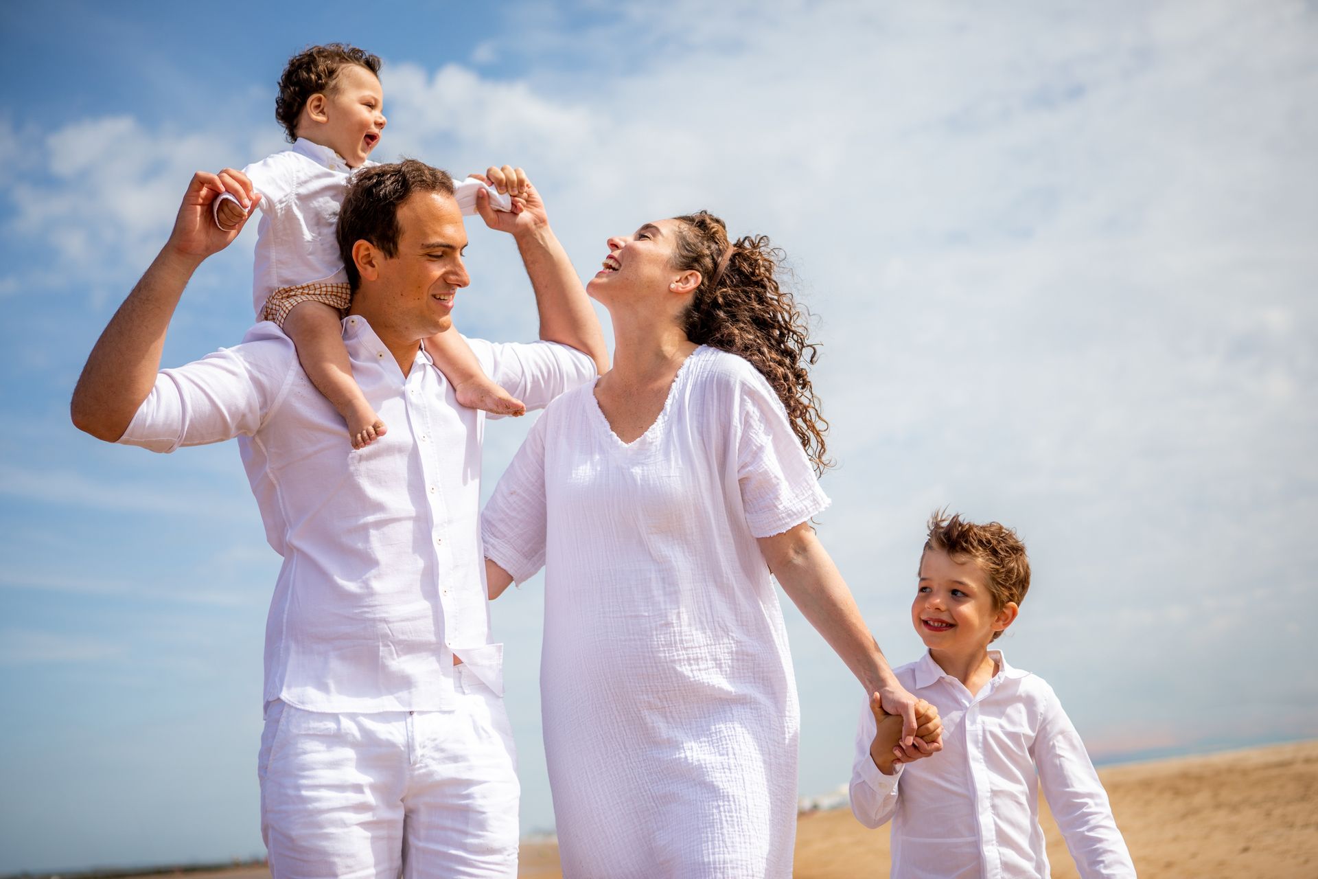 Een gezin van vier in witte kleding op het strand, lachend. Eén kind op de schouders, het andere in de hand. Blauwe lucht op de achtergrond.