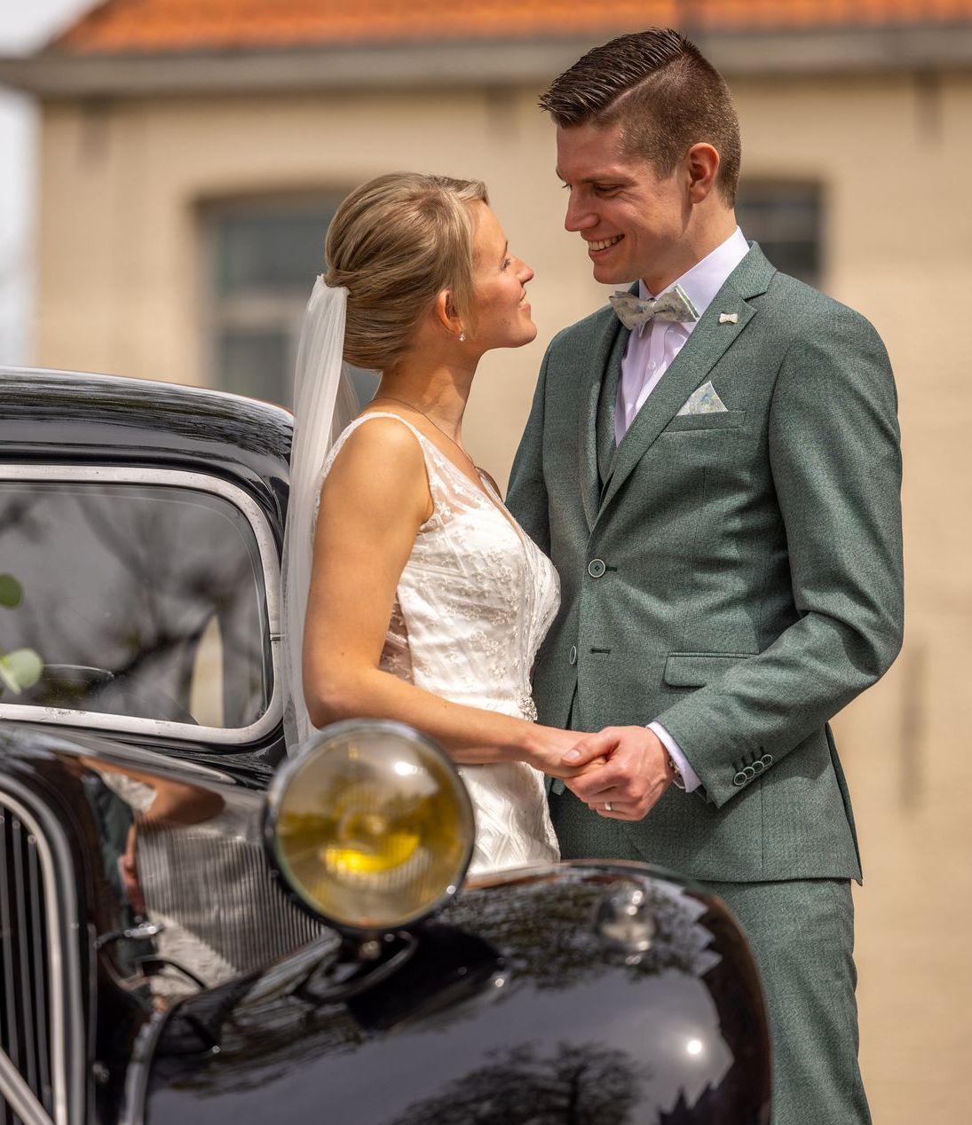 Het bruidspaar glimlacht naar elkaar, handen ineengeslagen. Hij draagt ​​een groen pak, zij een witte jurk, staande bij een klassieke auto.