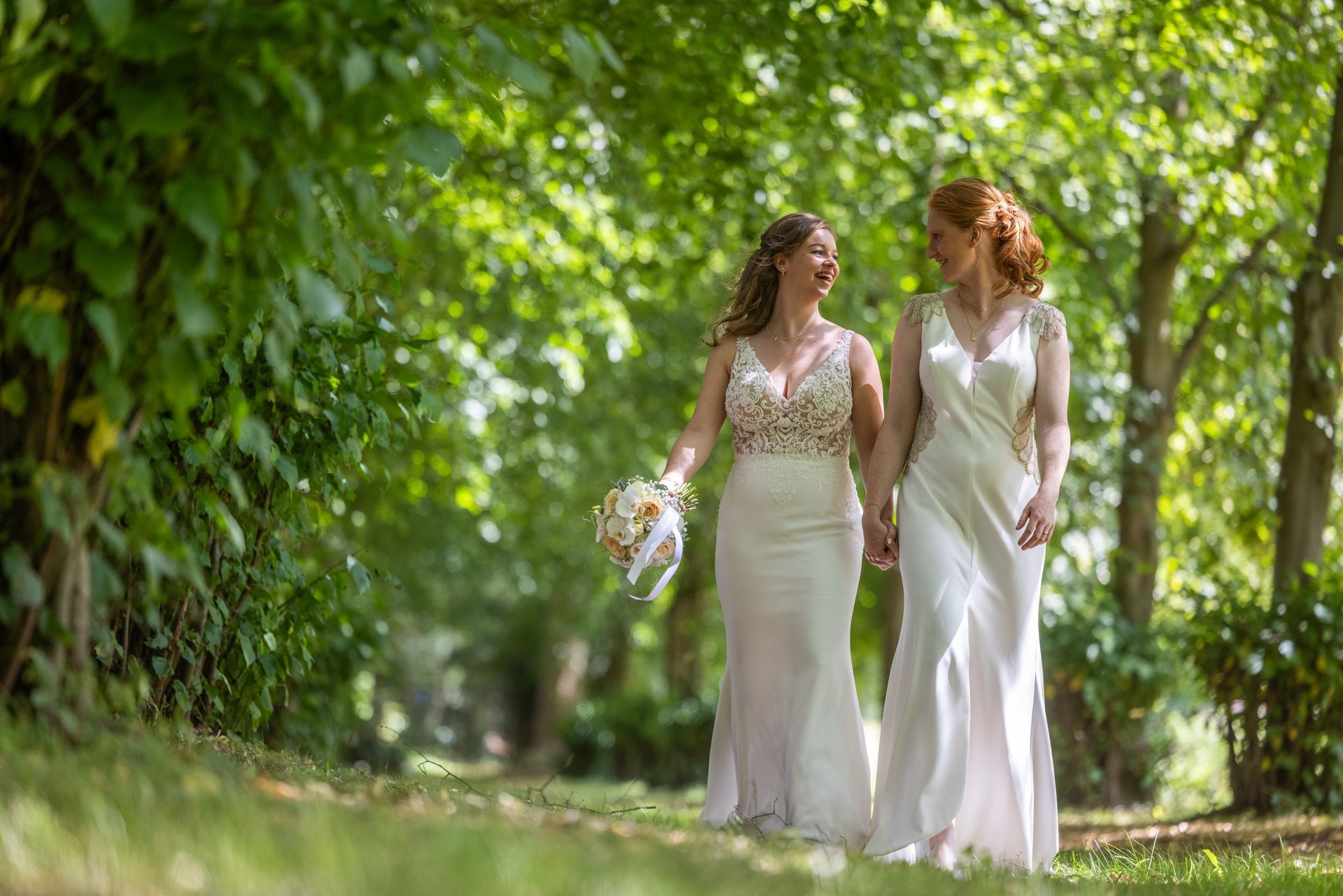 Twee bruiden in witte jurken lopen hand in hand over een pad, omgeven door bomen.