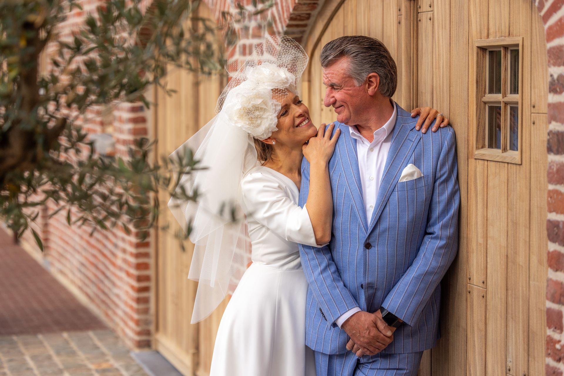 Bruid in witte jurk en bruidegom in blauw pak omhelzen elkaar, glimlachend, leunend tegen een houten deur, met een bakstenen muur op de achtergrond.