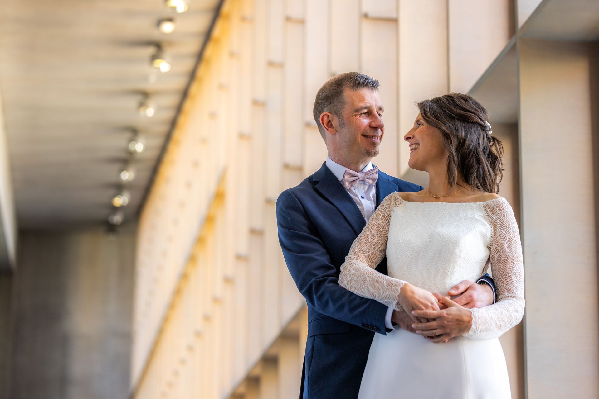 Een paar omhelst elkaar lachend bij een raam. De man draagt ​​een blauw pak, de vrouw een witte jurk.