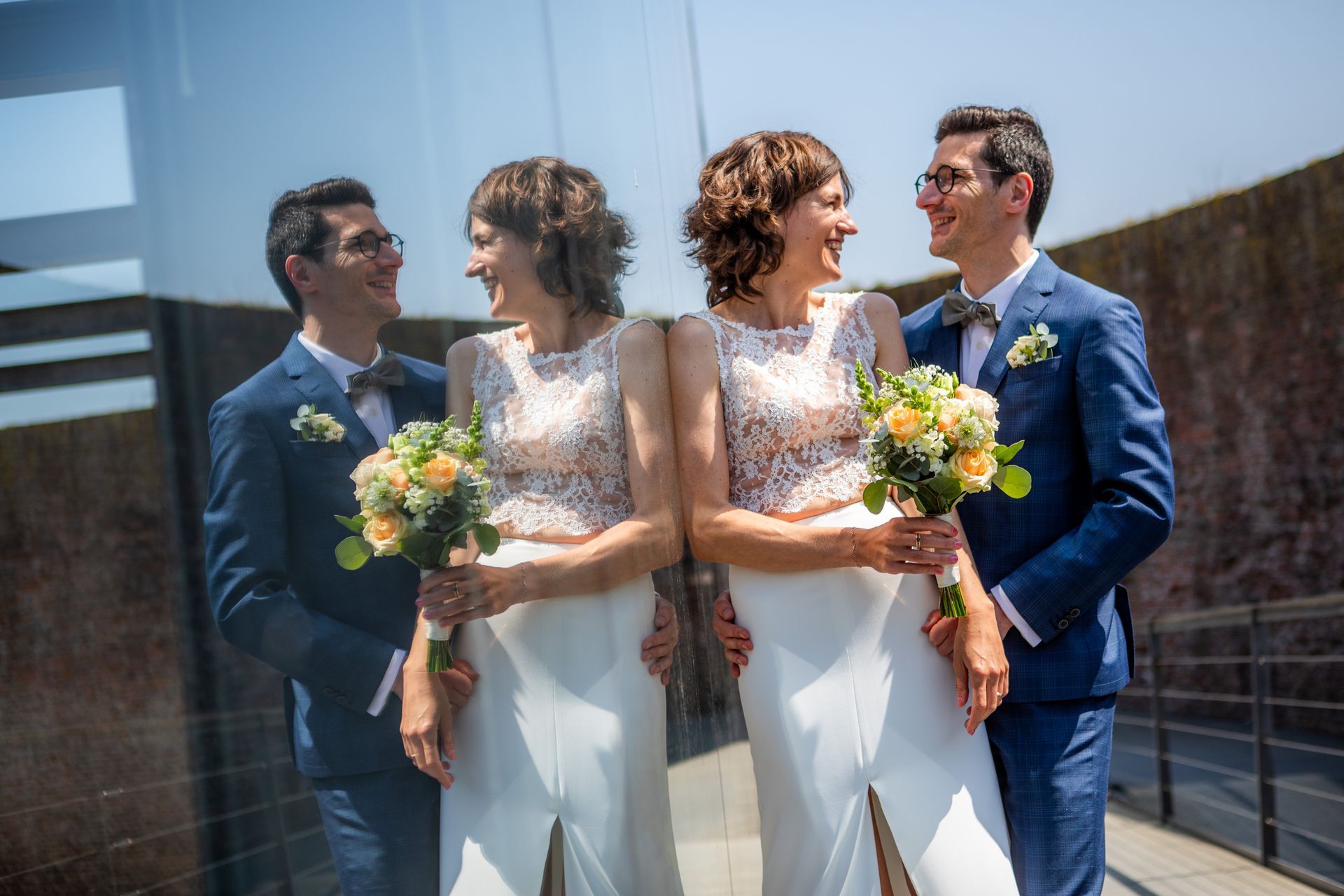 Bruid en bruidegom in bruidskleding, lachend en met een boeket in de hand, staand bij een spiegelend oppervlak.