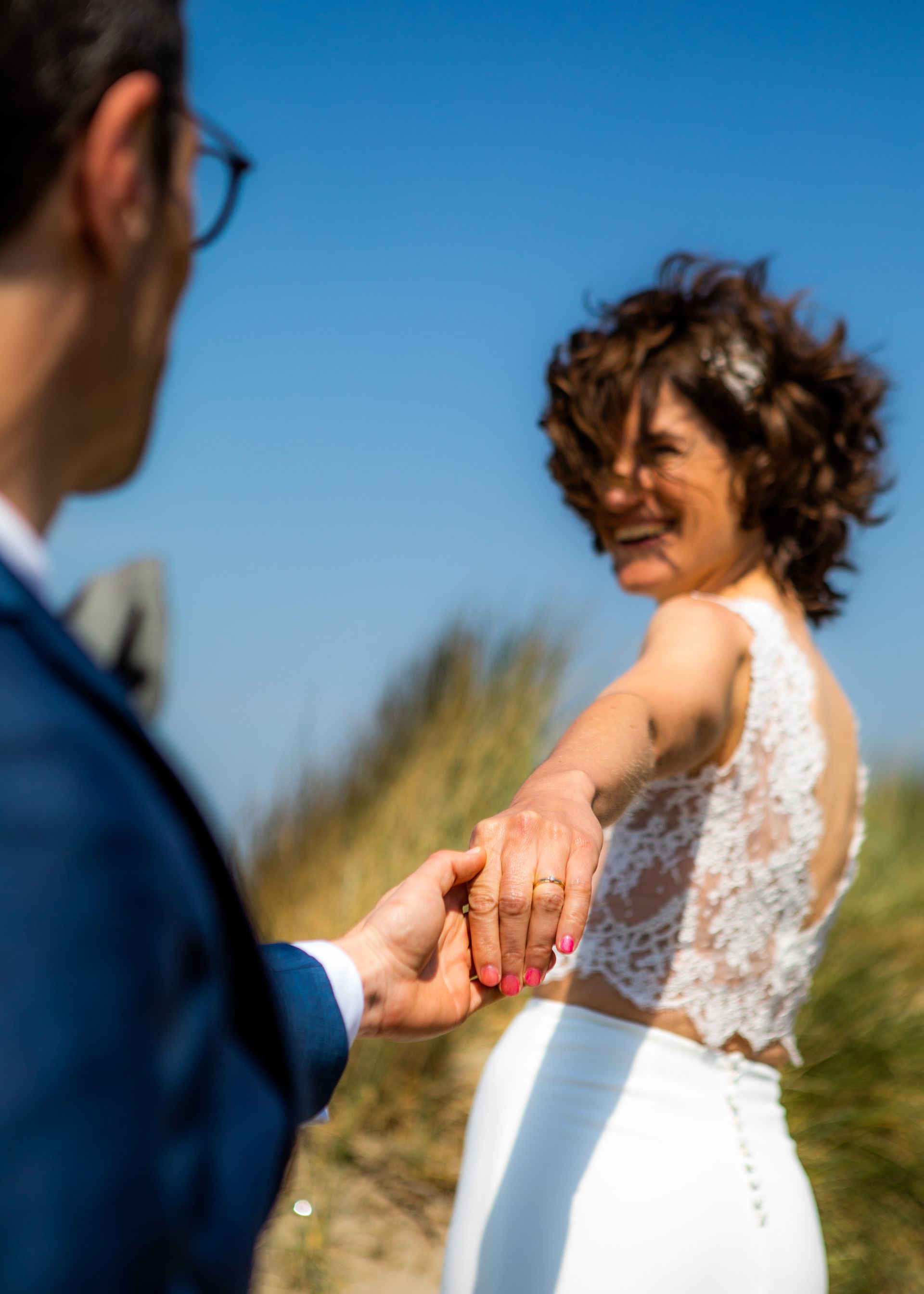 Een bruid in een kanten top en een witte rok lacht, terwijl ze op een zonnige dag hand in hand loopt met een man in een blauw pak.