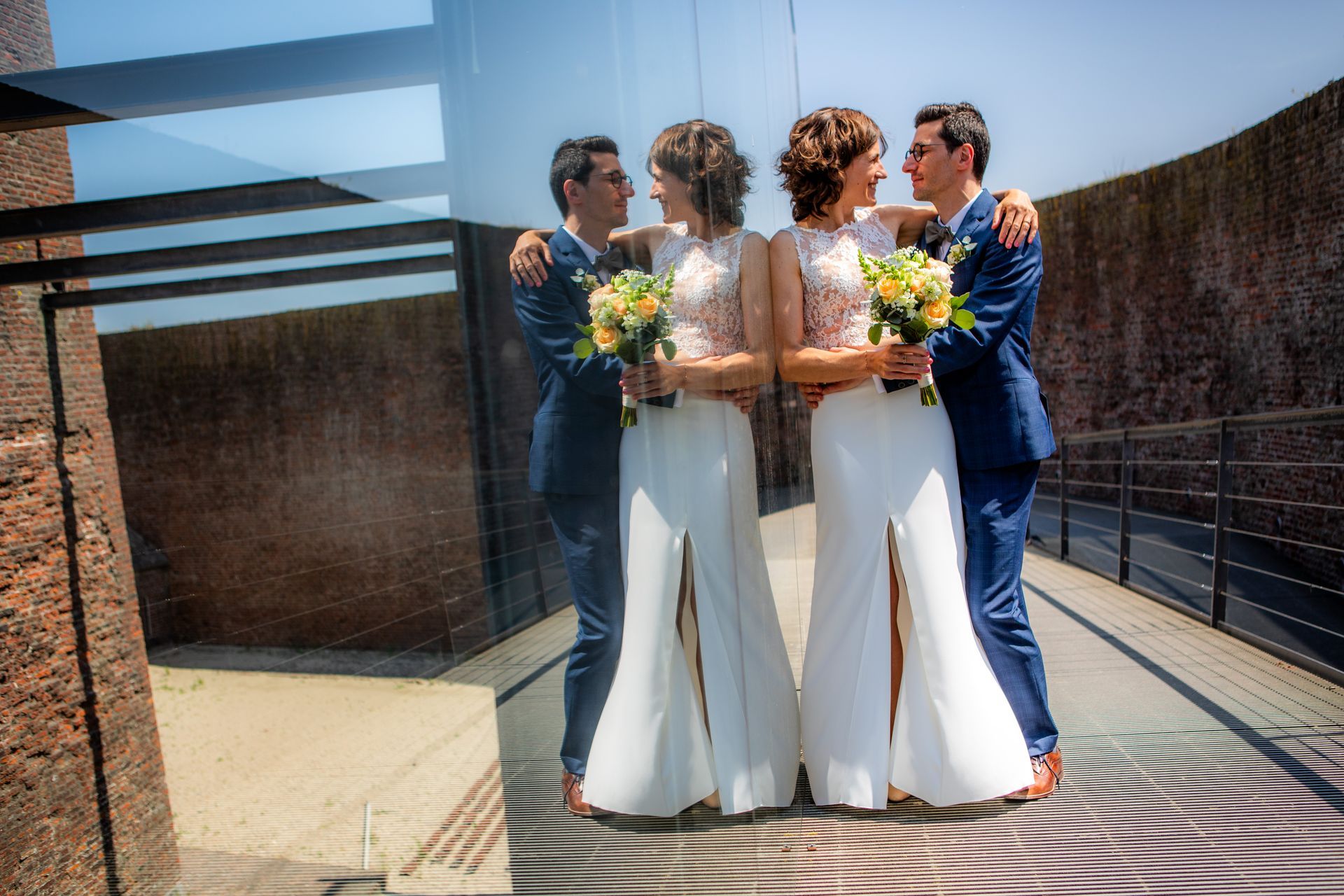 Een paar omhelst elkaar, lachend, met boeketten in de hand, weerspiegeld in een glazen wand. Zonnig buitenterras.