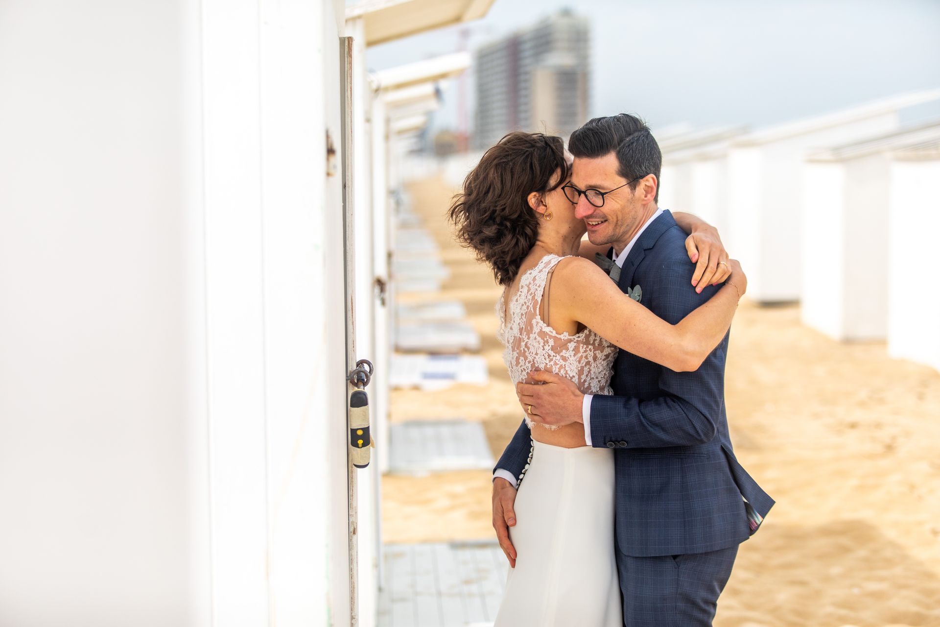 Een stel omhelst elkaar innig op een zandstrand, met witte cabana's op de achtergrond. Vrouw in witte jurk, man in blauw pak.