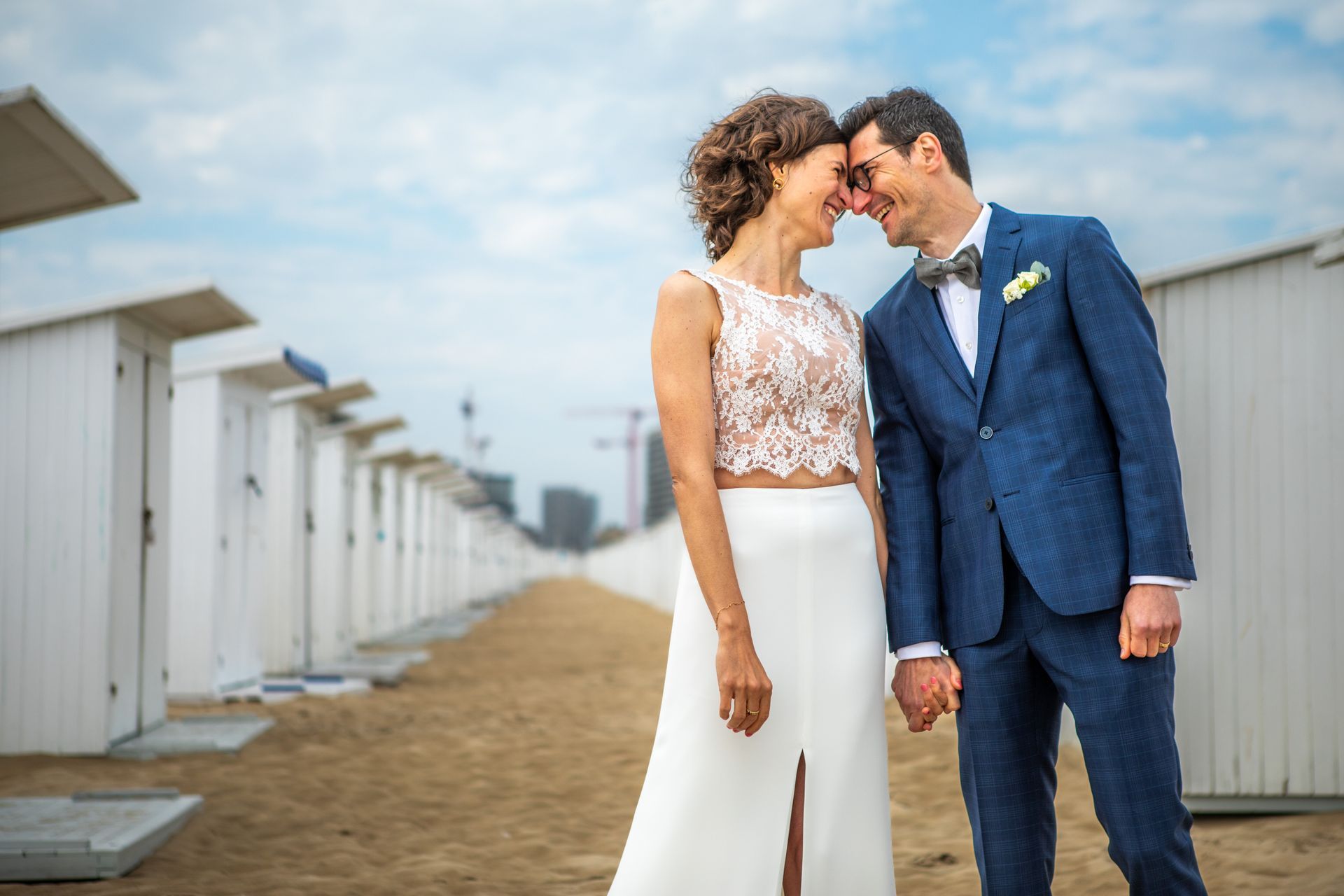 Een bruidspaar glimlacht en raakt elkaars voorhoofden aan op een zandstrand met witte strandhutten.