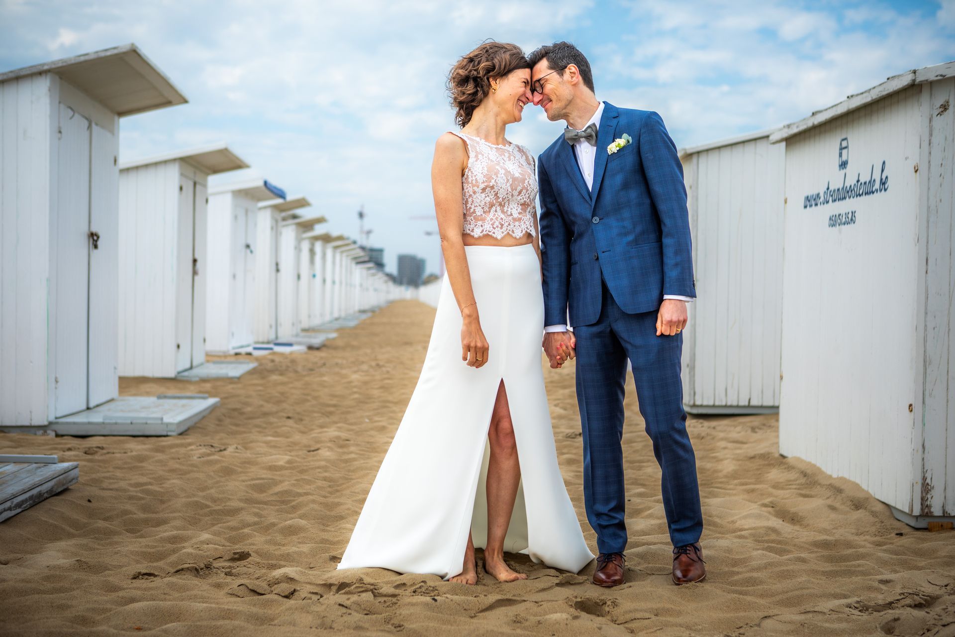 Bruid en bruidegom lachen, houden elkaars hand vast en raken elkaars voorhoofd aan op een zandstrand met witte strandhutten.