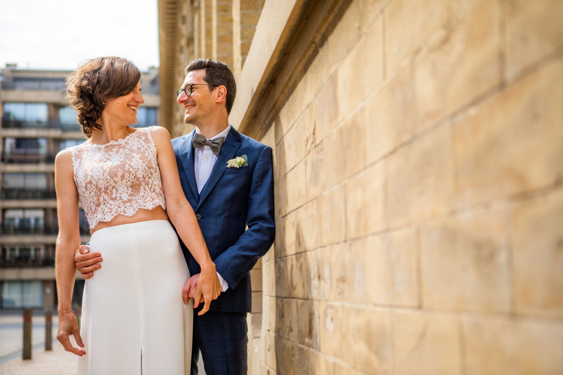 Het pasgetrouwde stel glimlacht naar elkaar, leunend tegen een bruine bakstenen muur. De bruid draagt ​​een kanten top en rok, de bruidegom een ​​blauw pak.