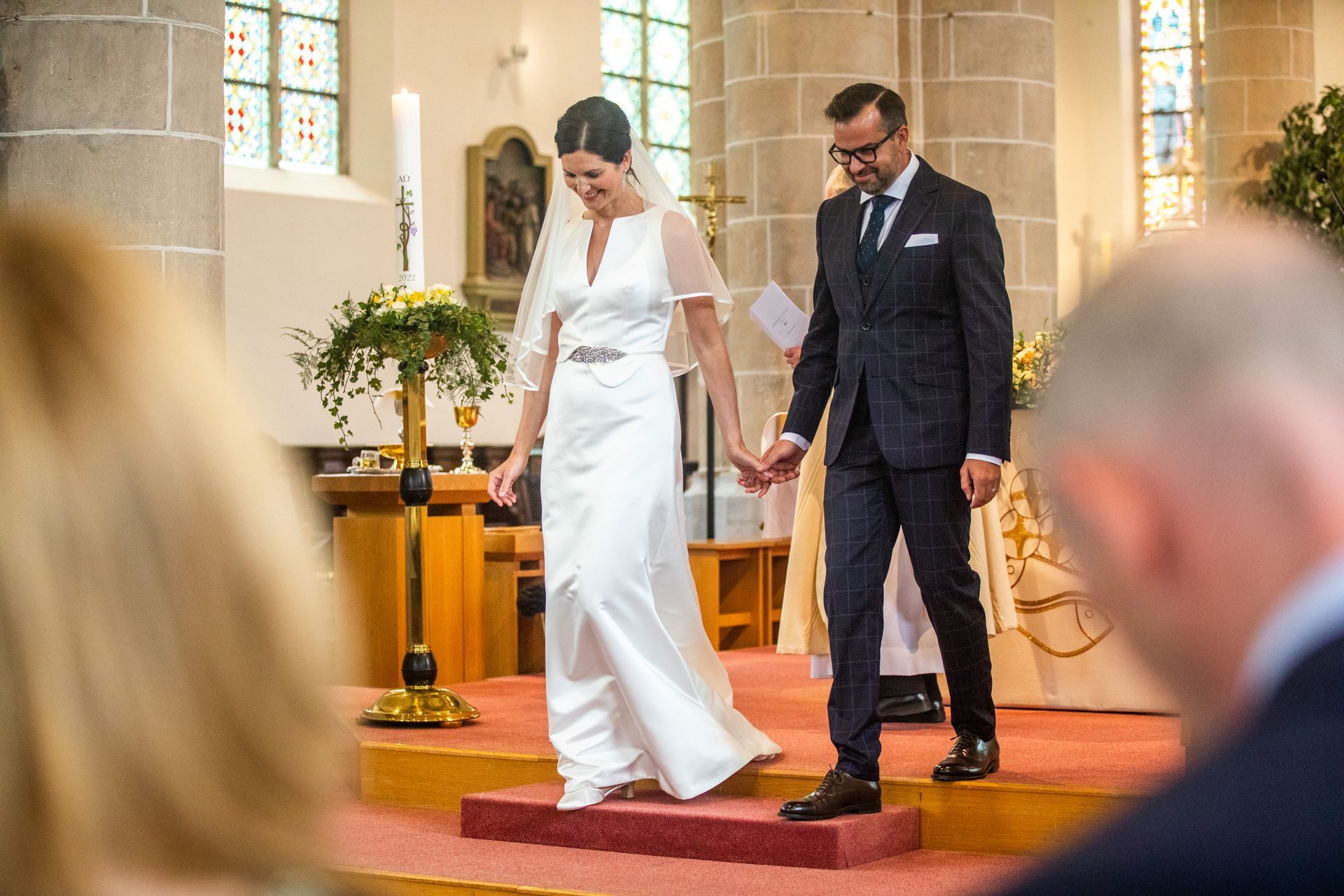 Het pasgetrouwde stel verlaat hand in hand de kerk. De bruid draagt ​​een witte jurk en sluier, de bruidegom een ​​donker pak.