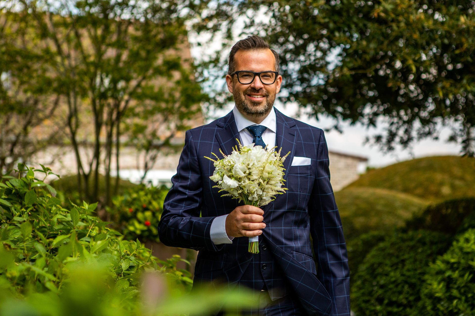 Man in blauw pak houdt wit boeket vast, lachend, in een tuin.