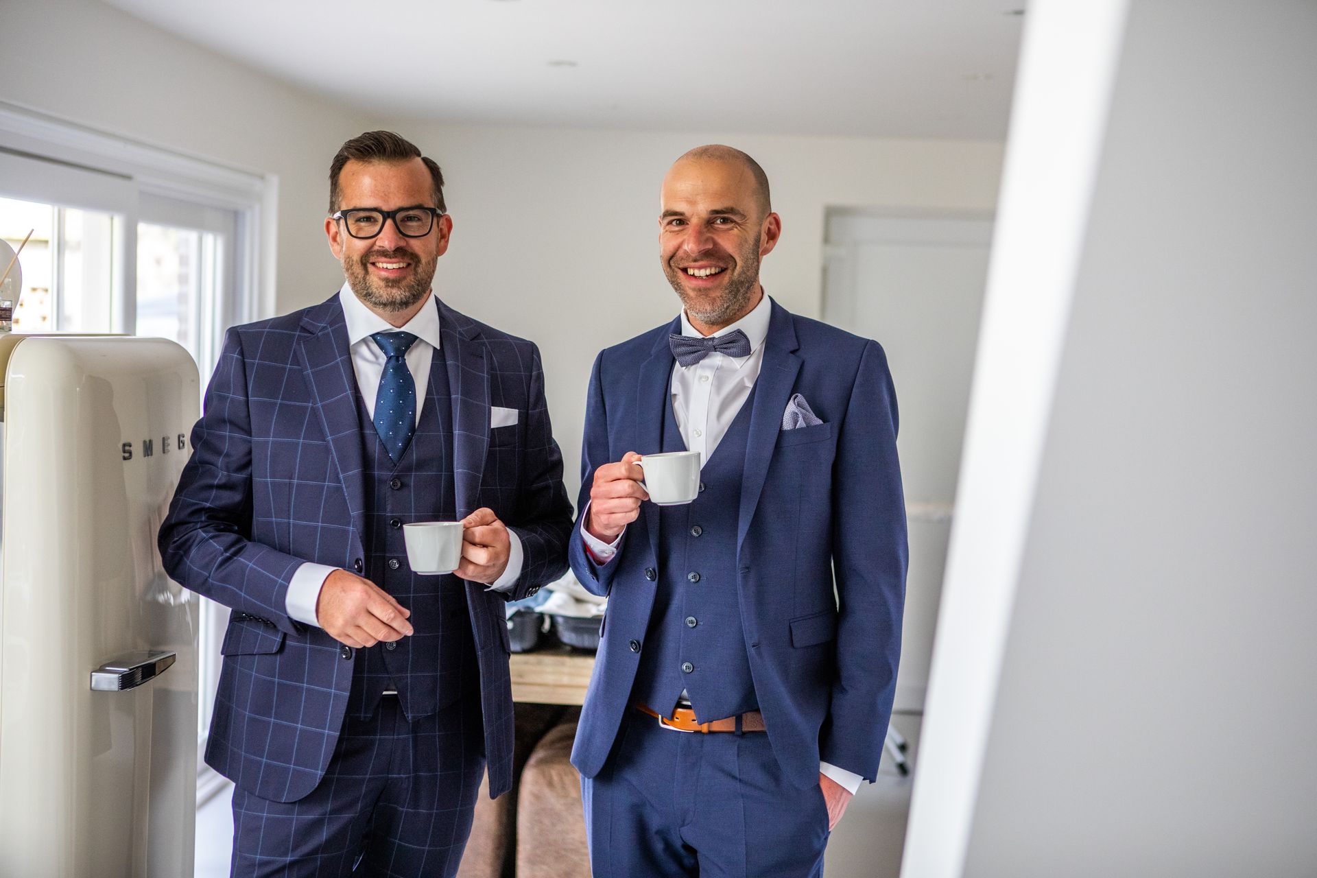 Twee mannen in blauwe pakken lachen, met een kop koffie in de hand, staan ​​in een felverlichte kamer.
