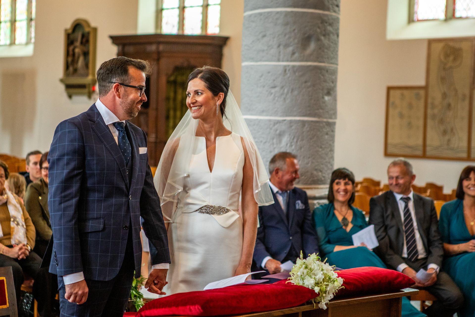 Bruid en bruidegom bij het altaar in de kerk, glimlachend naar elkaar. Gasten zitten. Zonlicht door de ramen.