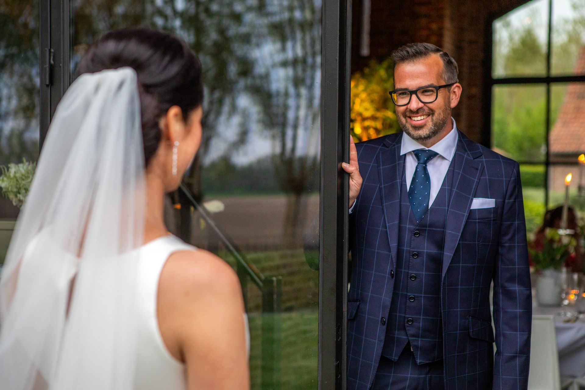 Bruid met sluier kijkt naar bruidegom in blauw pak die glimlachend bij de deuropening staat.