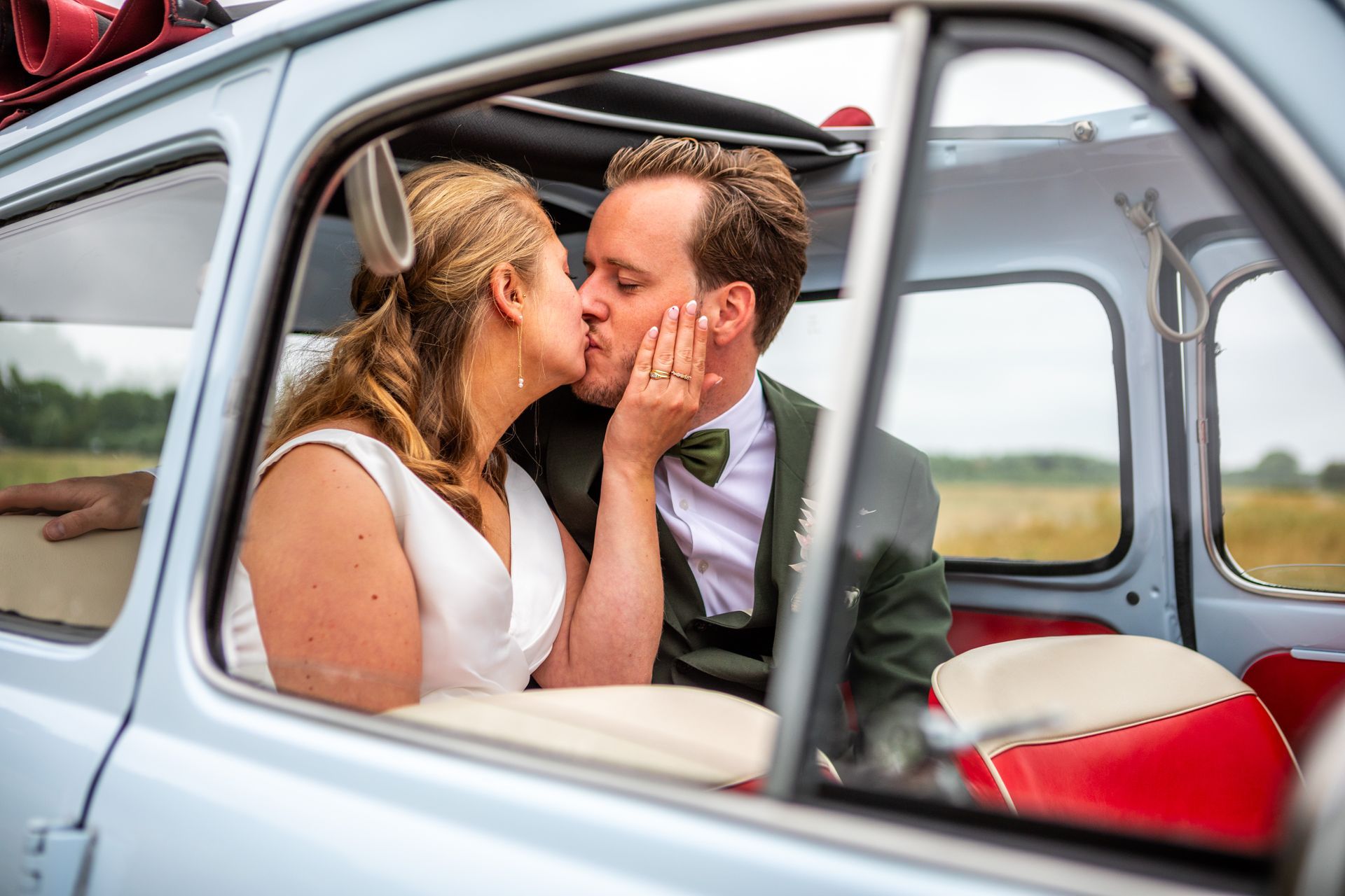 Bruid en bruidegom kussen elkaar in een blauwe vintage auto; interieur rood en wit.