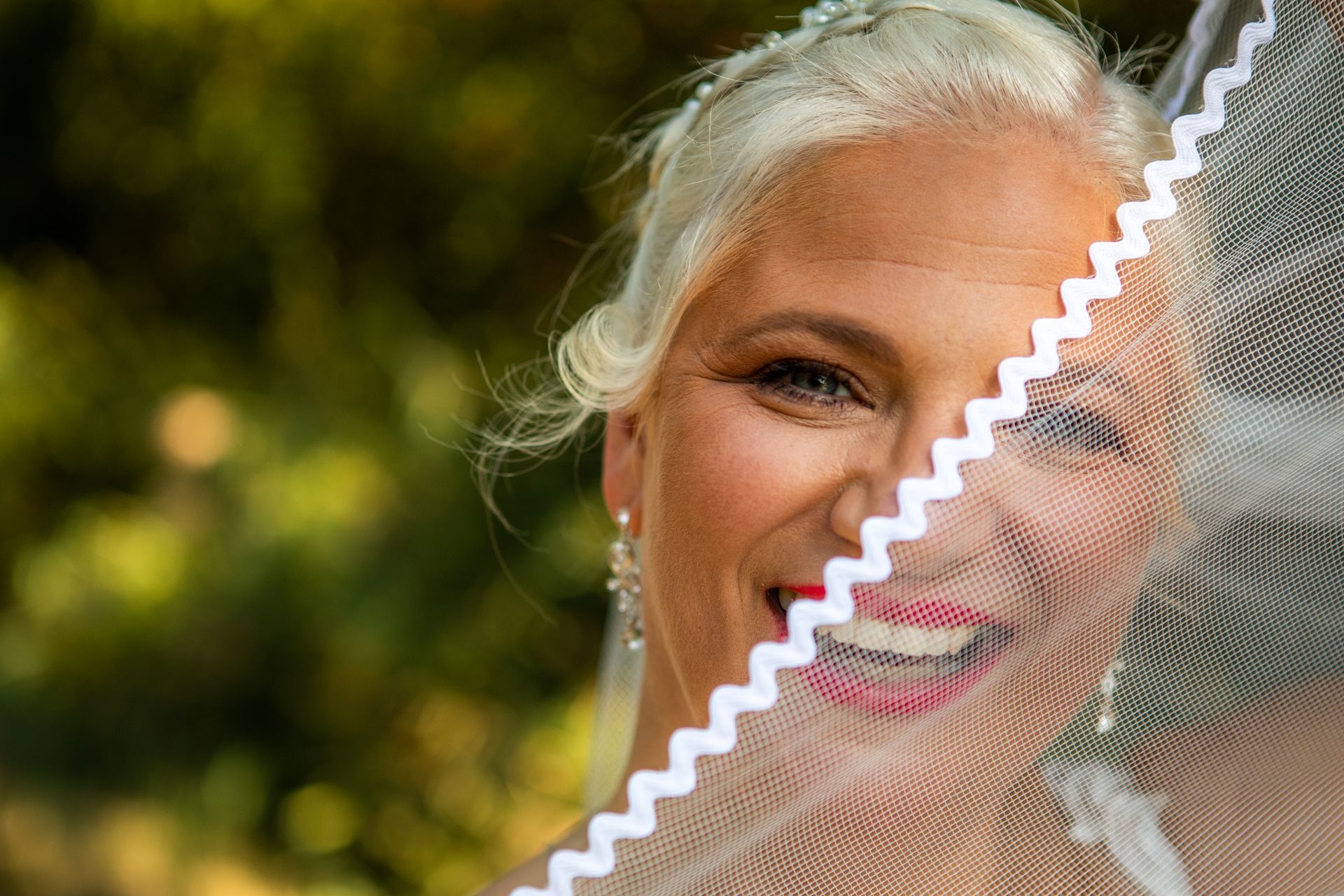 Bruid met blond haar, glimlachend, glurend door een witte sluier met een zigzagrand, buiten.