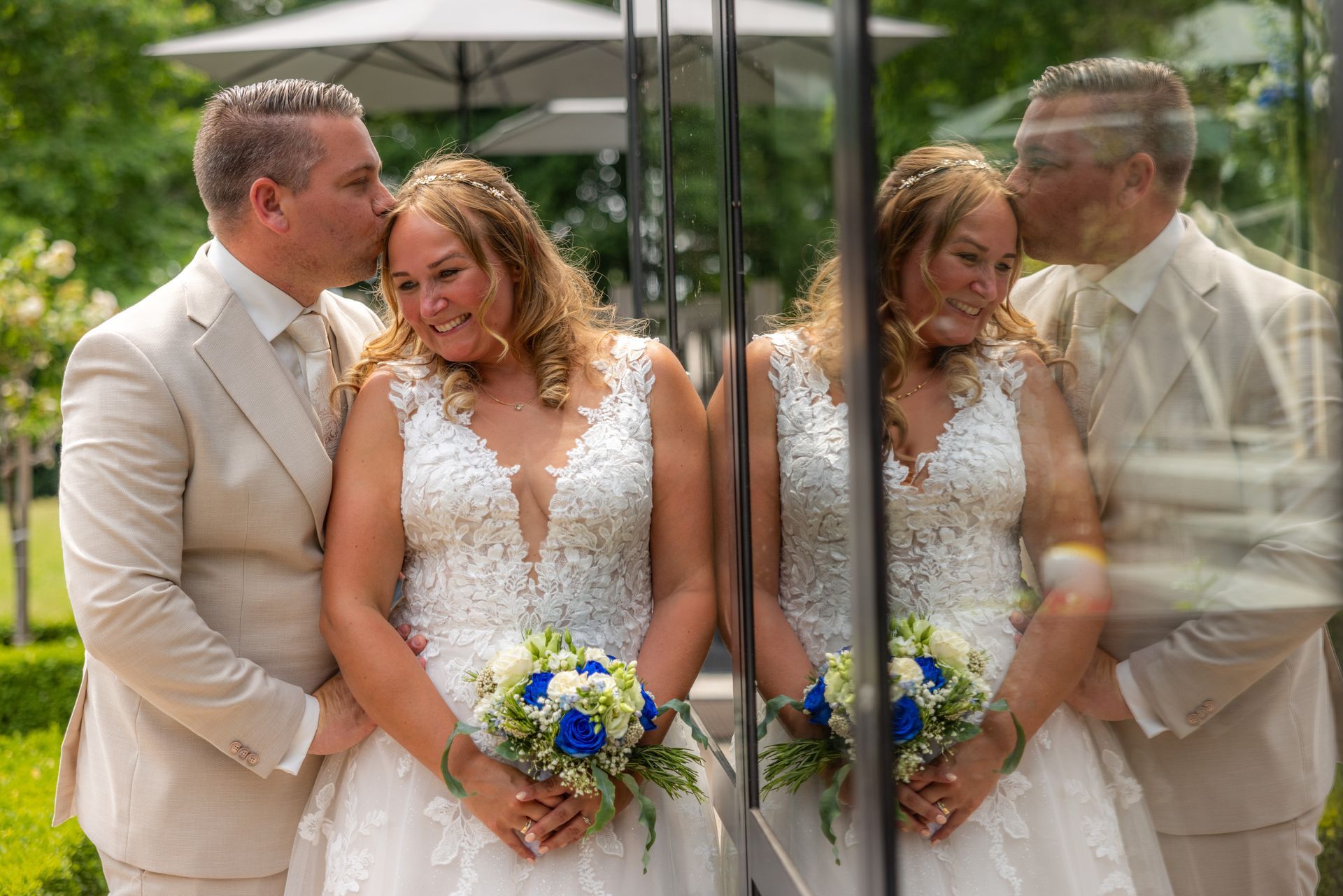 De bruidegom kust het voorhoofd van de bruid, beiden glimlachend, weerspiegeld in een raam. De bruid houdt een blauw-wit boeket vast; ze staan ​​buiten.
