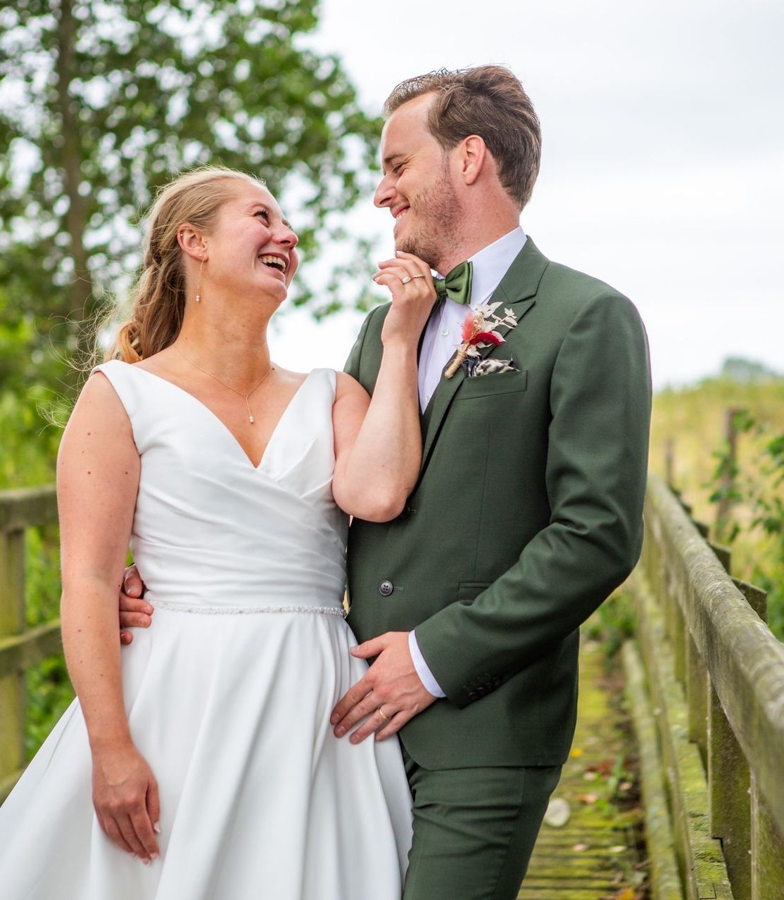 Bruid en bruidegom lachen op een houten brug, man in een groen pak, vrouw in een witte jurk, buitenomgeving.