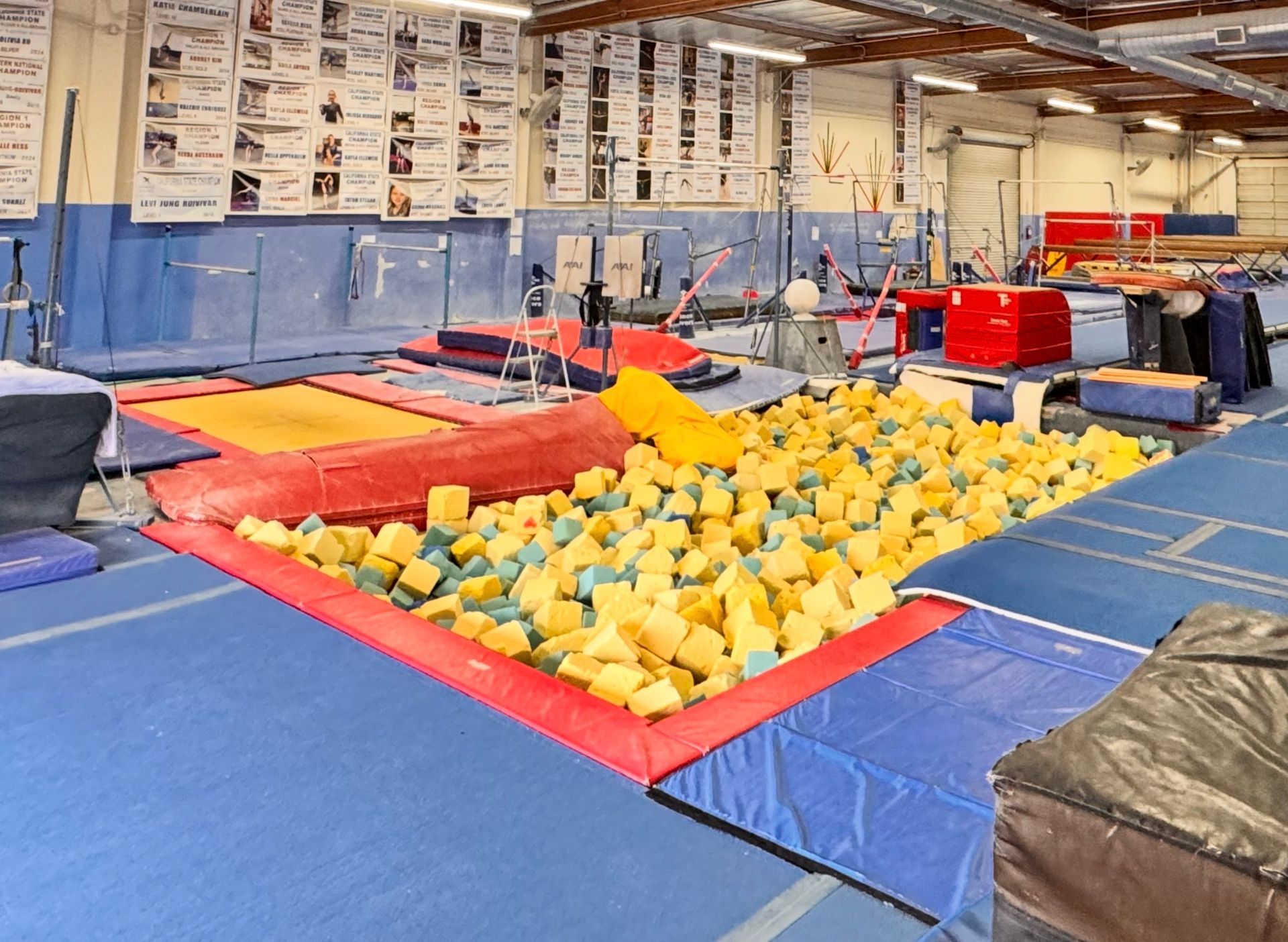 Gymnasium interior with gymnastics equipment, including a foam pit, uneven bars, and mats.