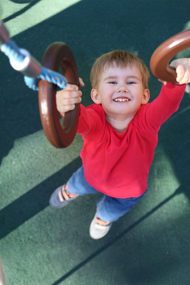 Boy in red shirt smiles, hanging from playground rings.