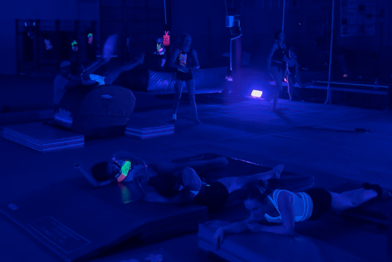 People on floor glowing in a dark room under blue light; aerial silks in background.