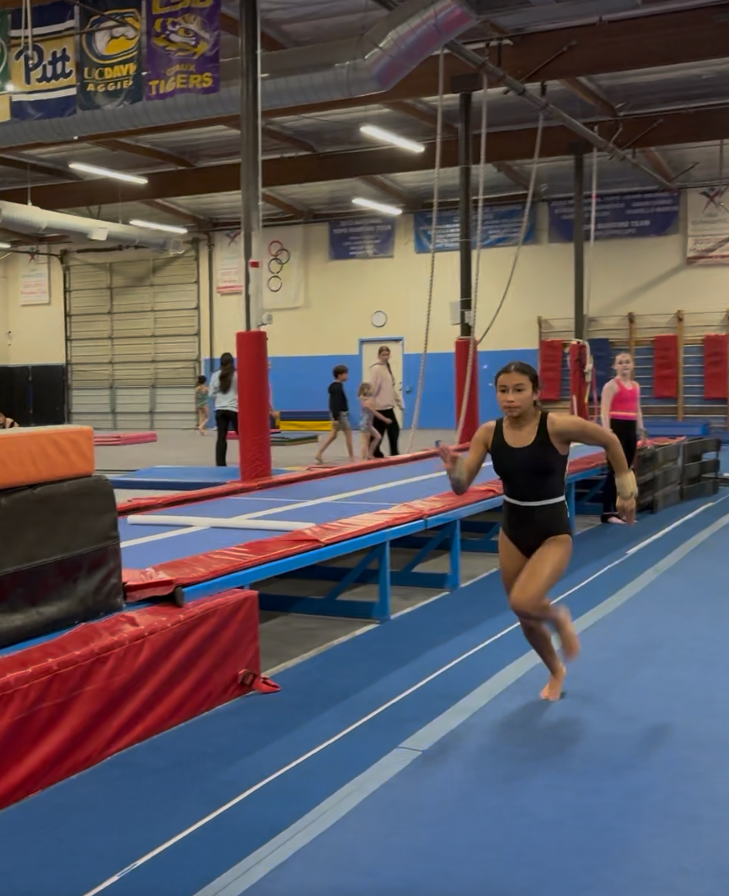 Gymnast running on a blue mat, wearing a black leotard. Gym setting with other people and equipment in the background.