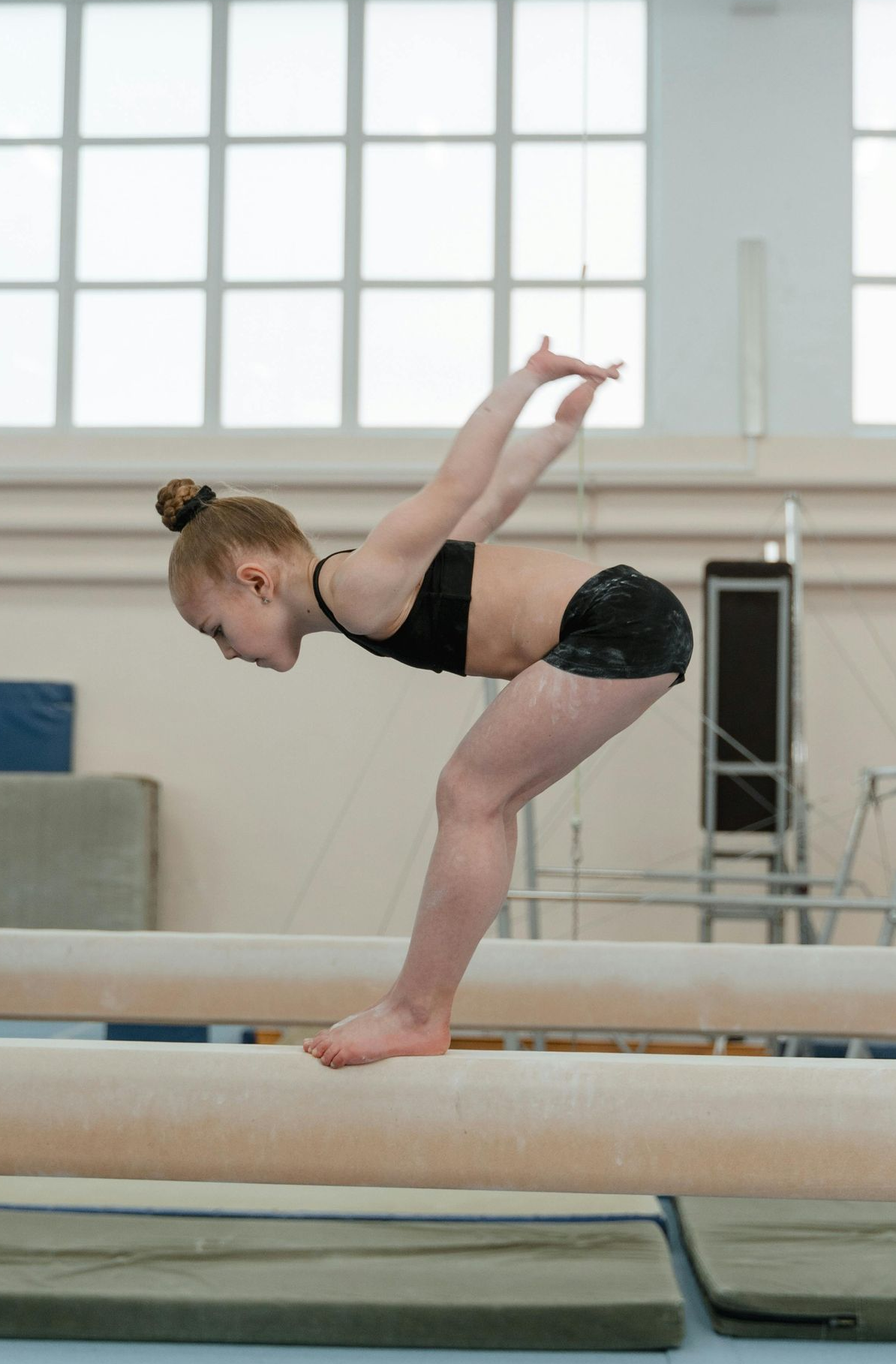 Gymnast on a balance beam, bending backward with arms raised, in a gym.