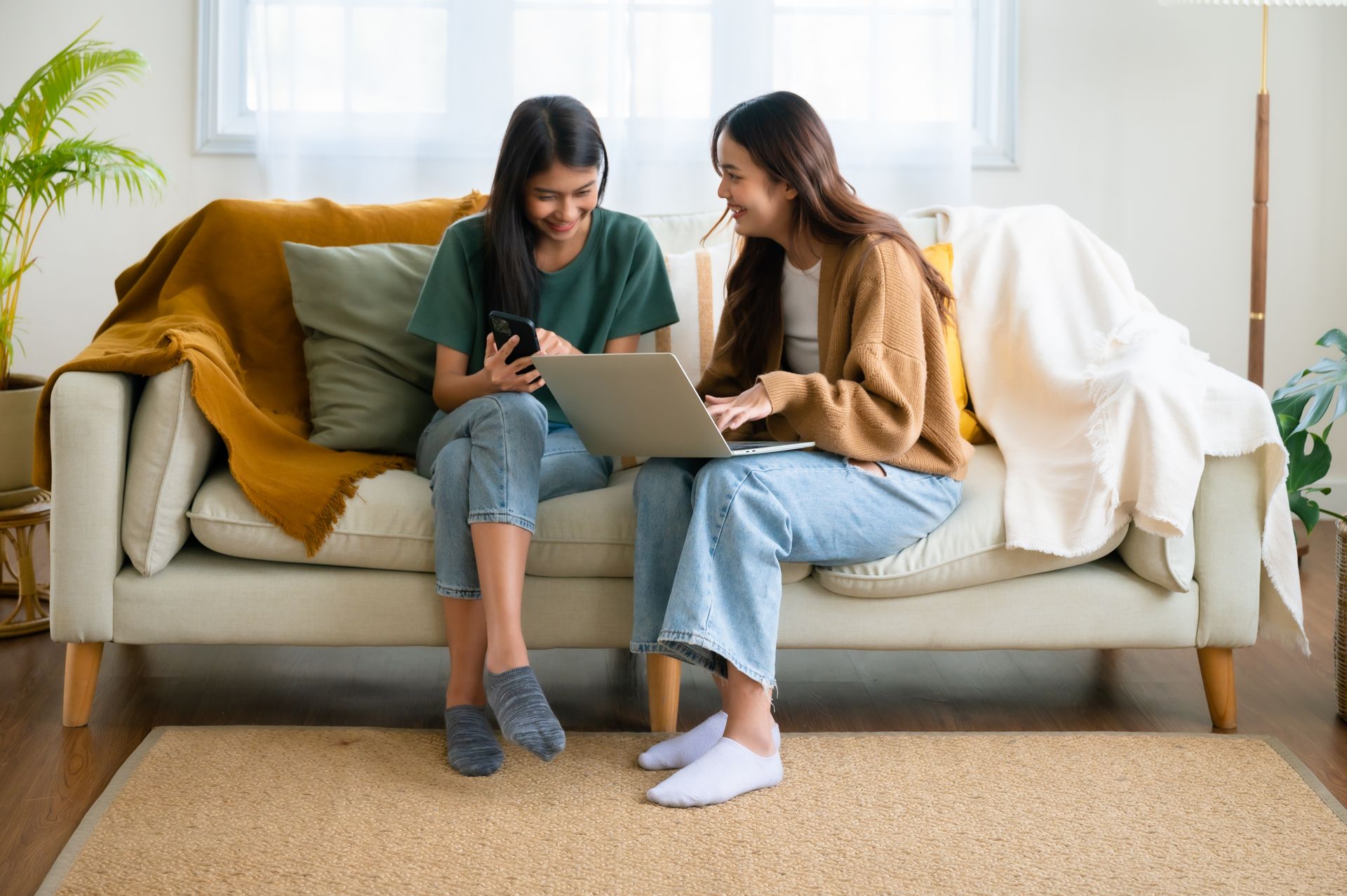 Two women are sitting on a couch looking at a laptop and a cell phone.