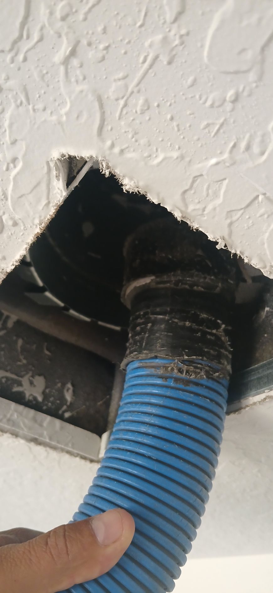A hand holds a blue corrugated hose connected to a black exhaust fan vent installed in a textured white ceiling.