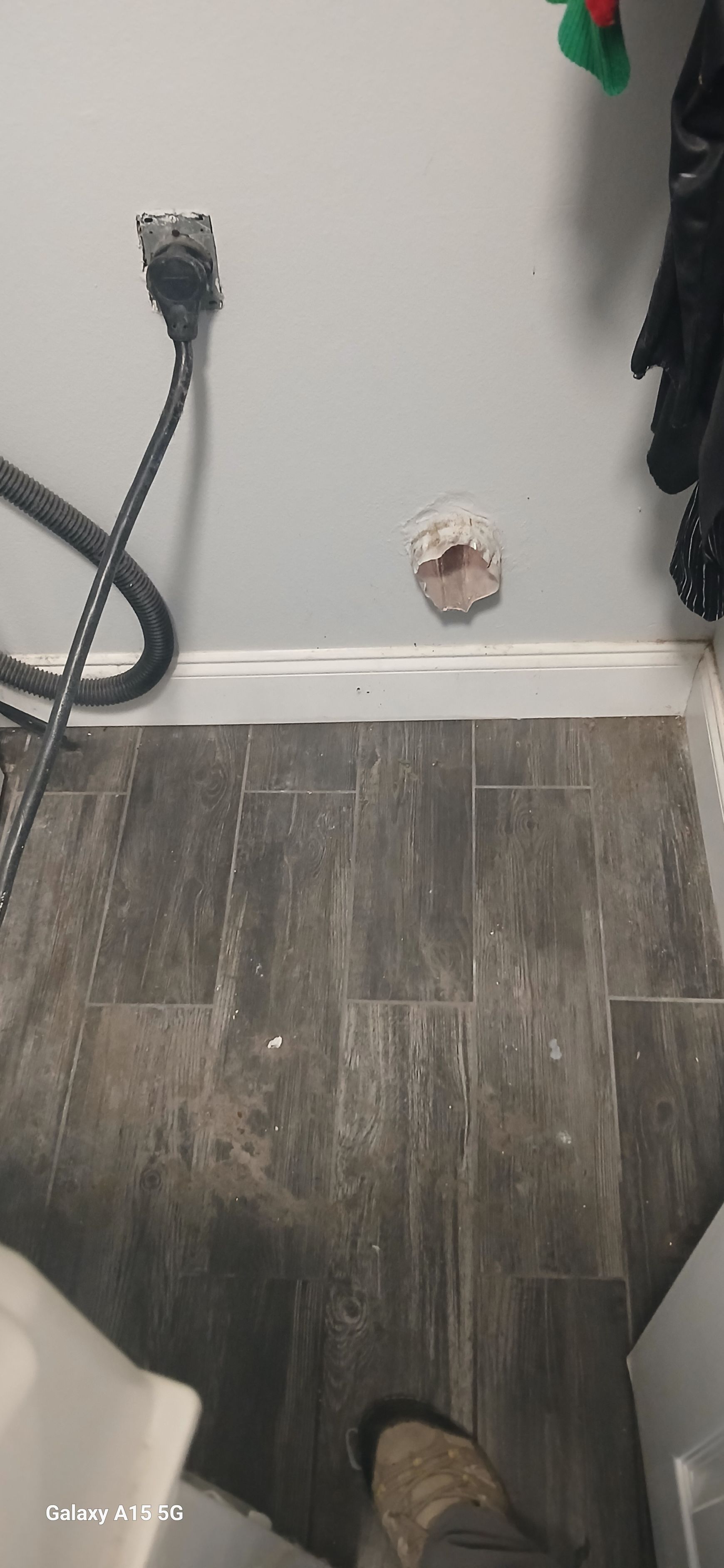 A wall-mounted dryer vent hole in a room with dark wood-look tile flooring and a nearby electrical outlet.