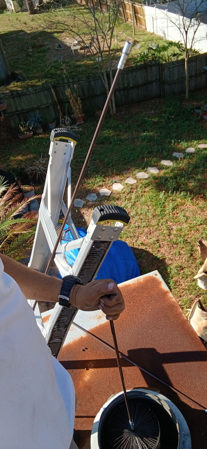 A person holds a long, thin rod extending from a container, with a white ladder and yard in the background.