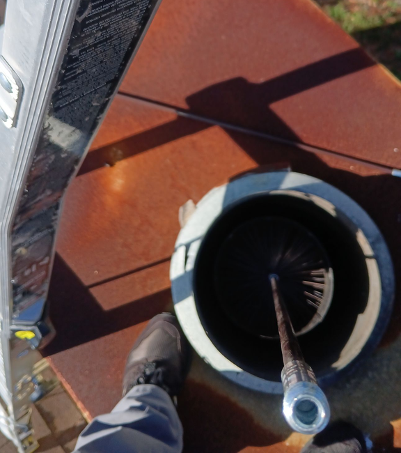 A person wearing a grey hoodie, knit cap, and yellow work gloves performs maintenance on a red brick chimney on a roof.