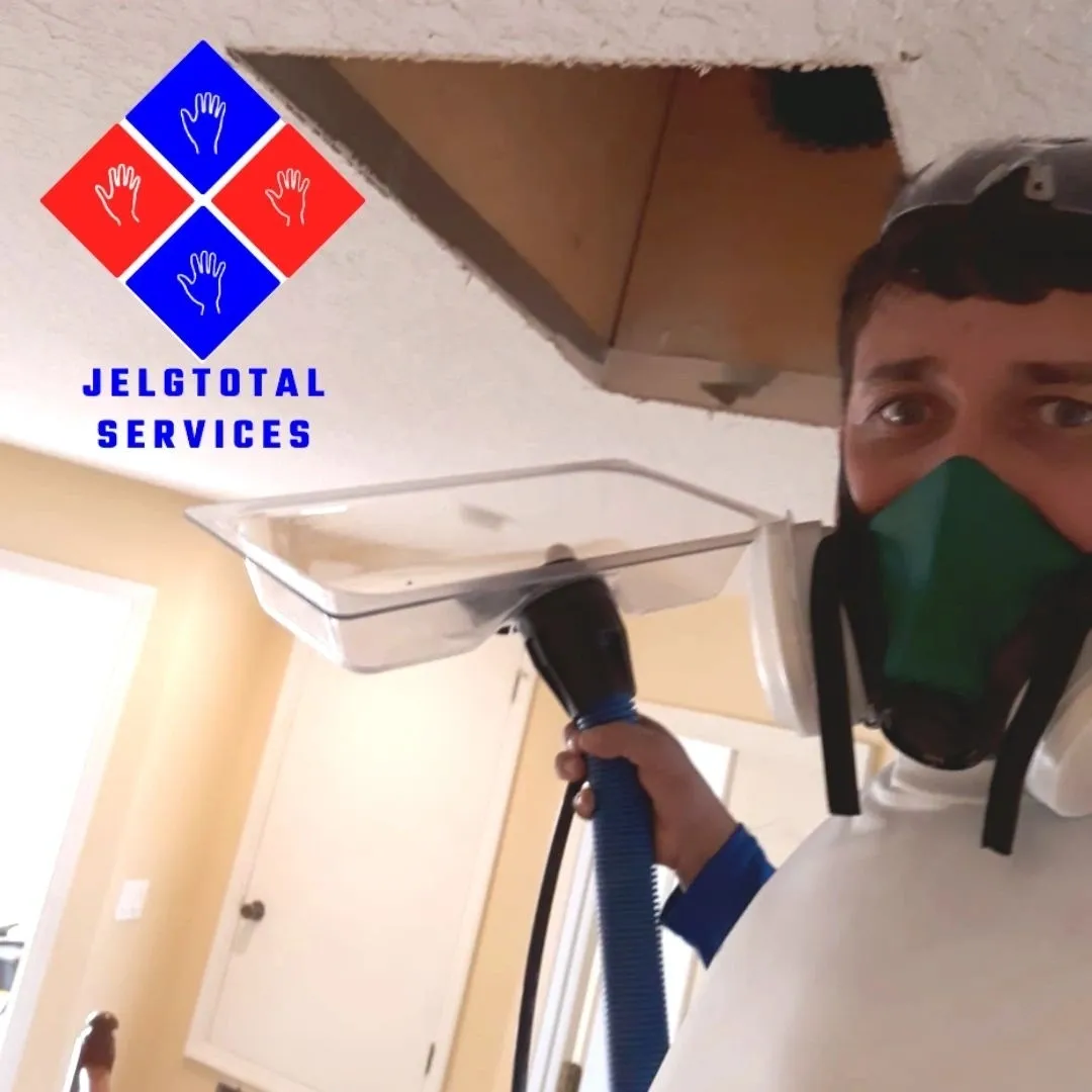 A worker wearing a protective mask and suit holds a vacuum attachment toward a ceiling opening in an indoor setting.