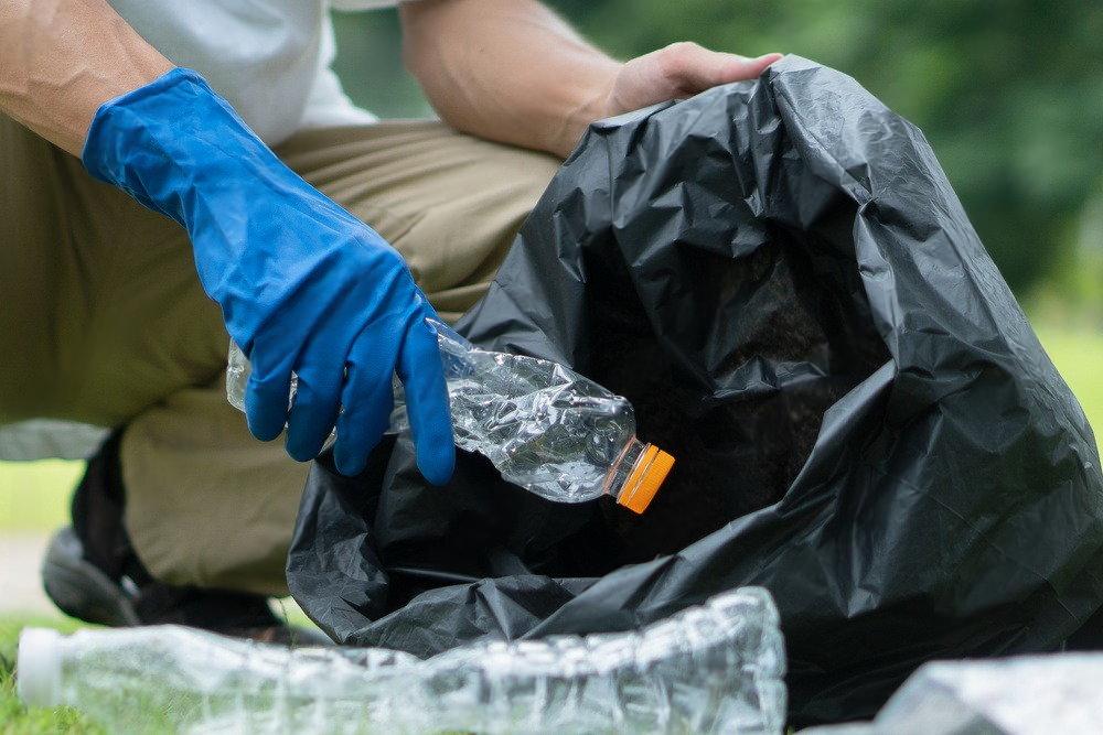 Person in blue gloves putting a plastic bottle into a black trash bag on a grassy area.