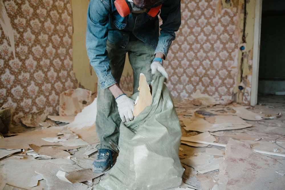 Person in a respirator putting debris into a bag during a room renovation; wallpaper and floor pieces strewn.