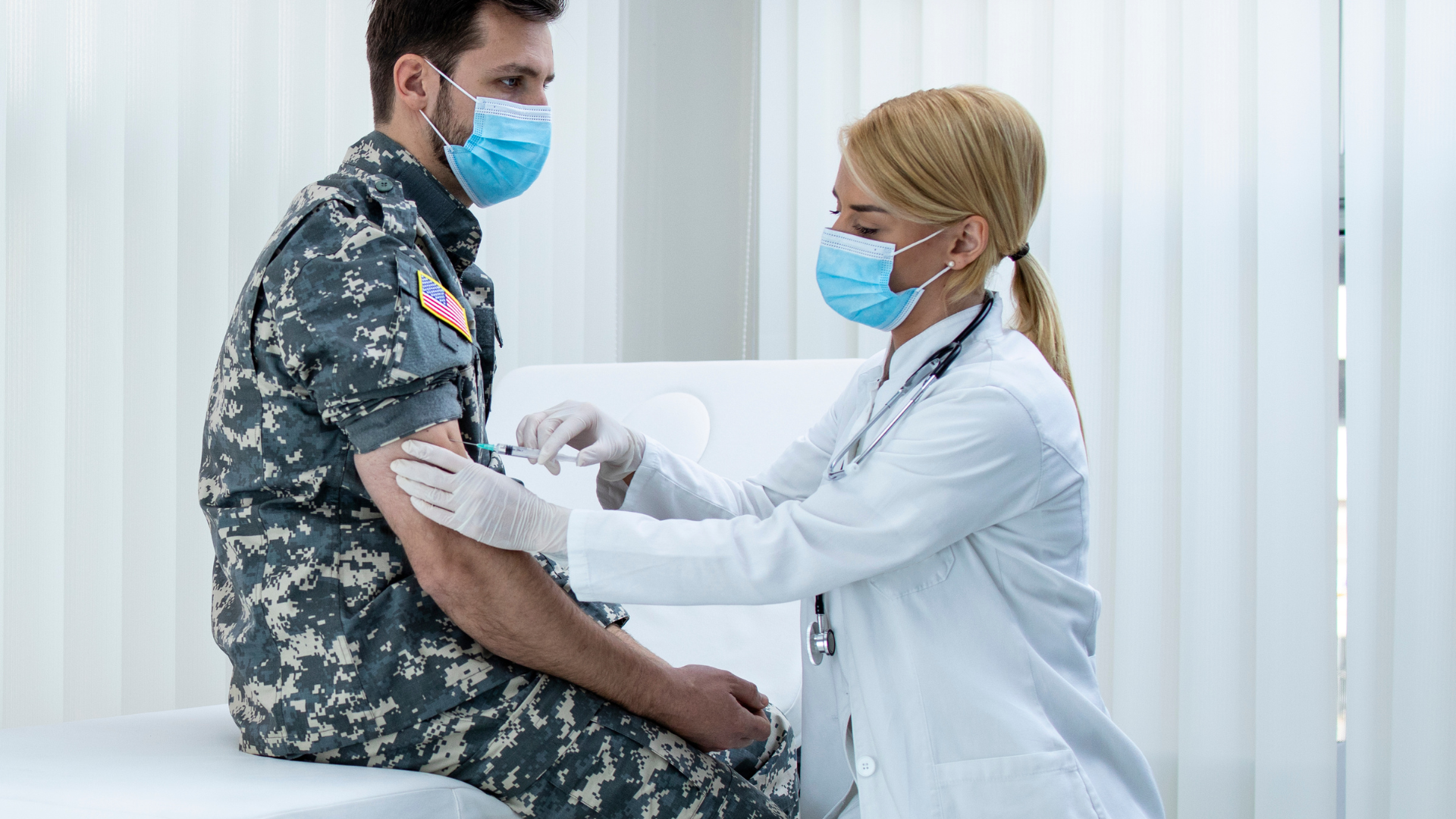 Older man in plaid shirt talking to person out of view, American flag in background.