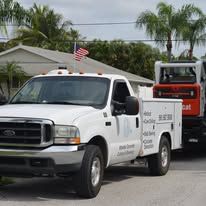 White utility truck with an American flag parked in a residential street, towing a red skid steer loader.