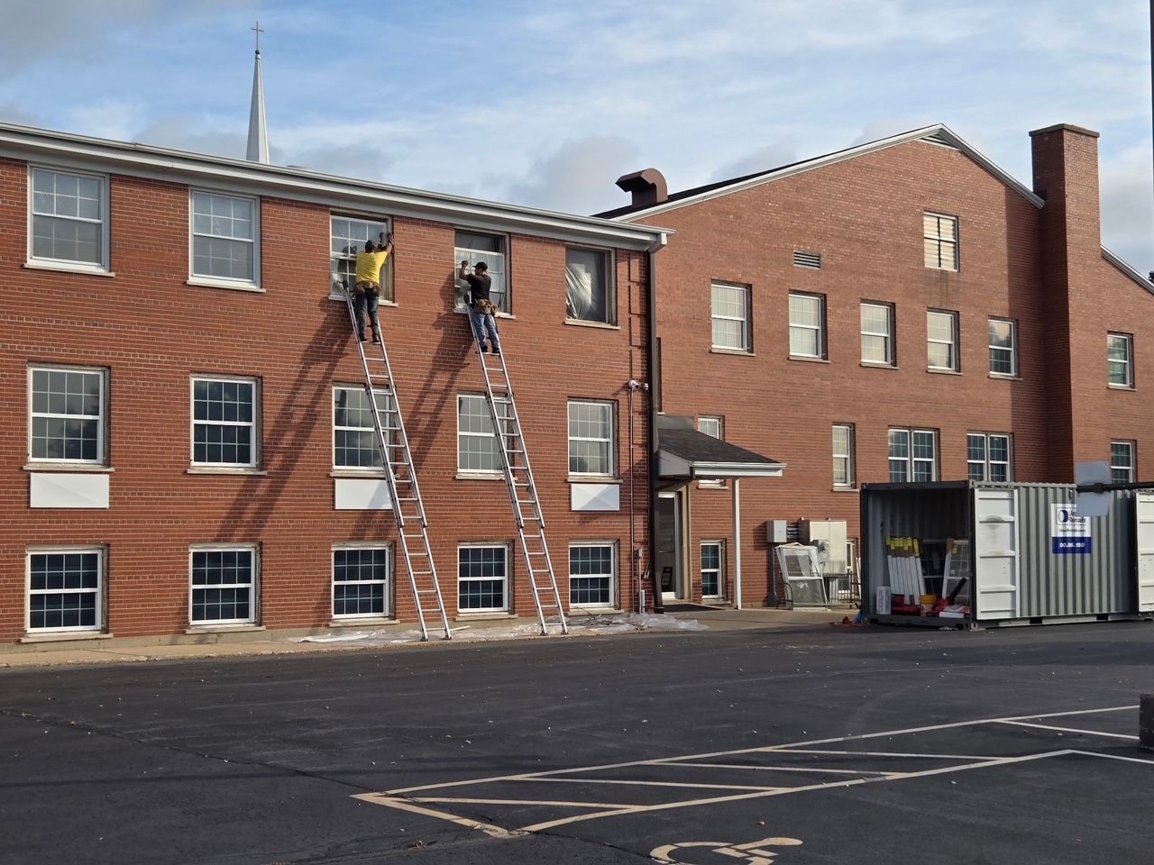 Two people on ladders installing windows of a brick building. Blue sky, a parking lot, and a storage container are visible.