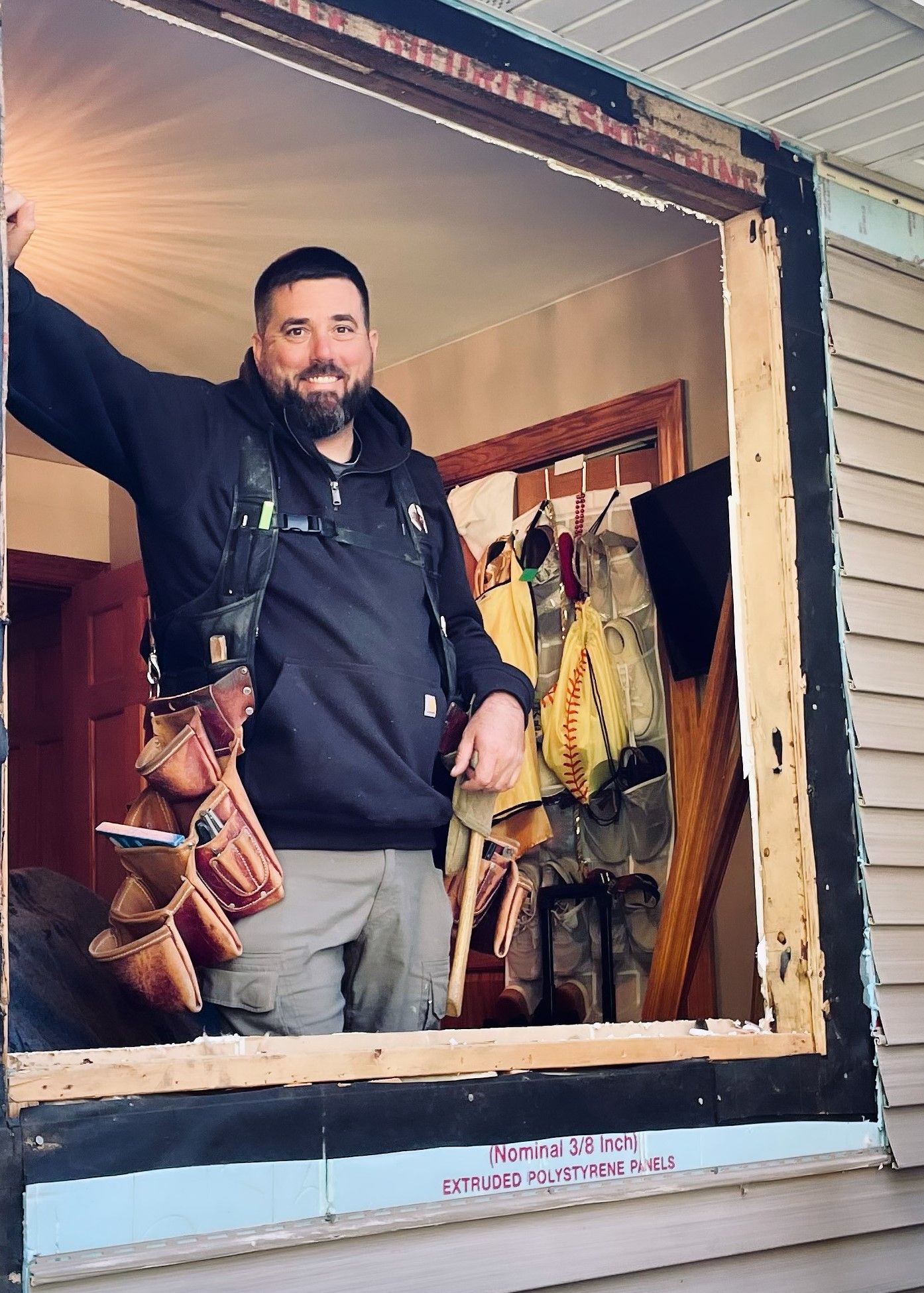 A man stands in an unfinished window frame wearing a tool belt, smiling. Outside is beige siding.
