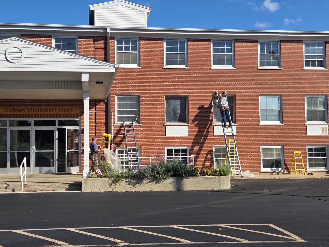 Two workers on ladders paint a brick building. Entrance with white awning and windows. Asphalt parking lot.
