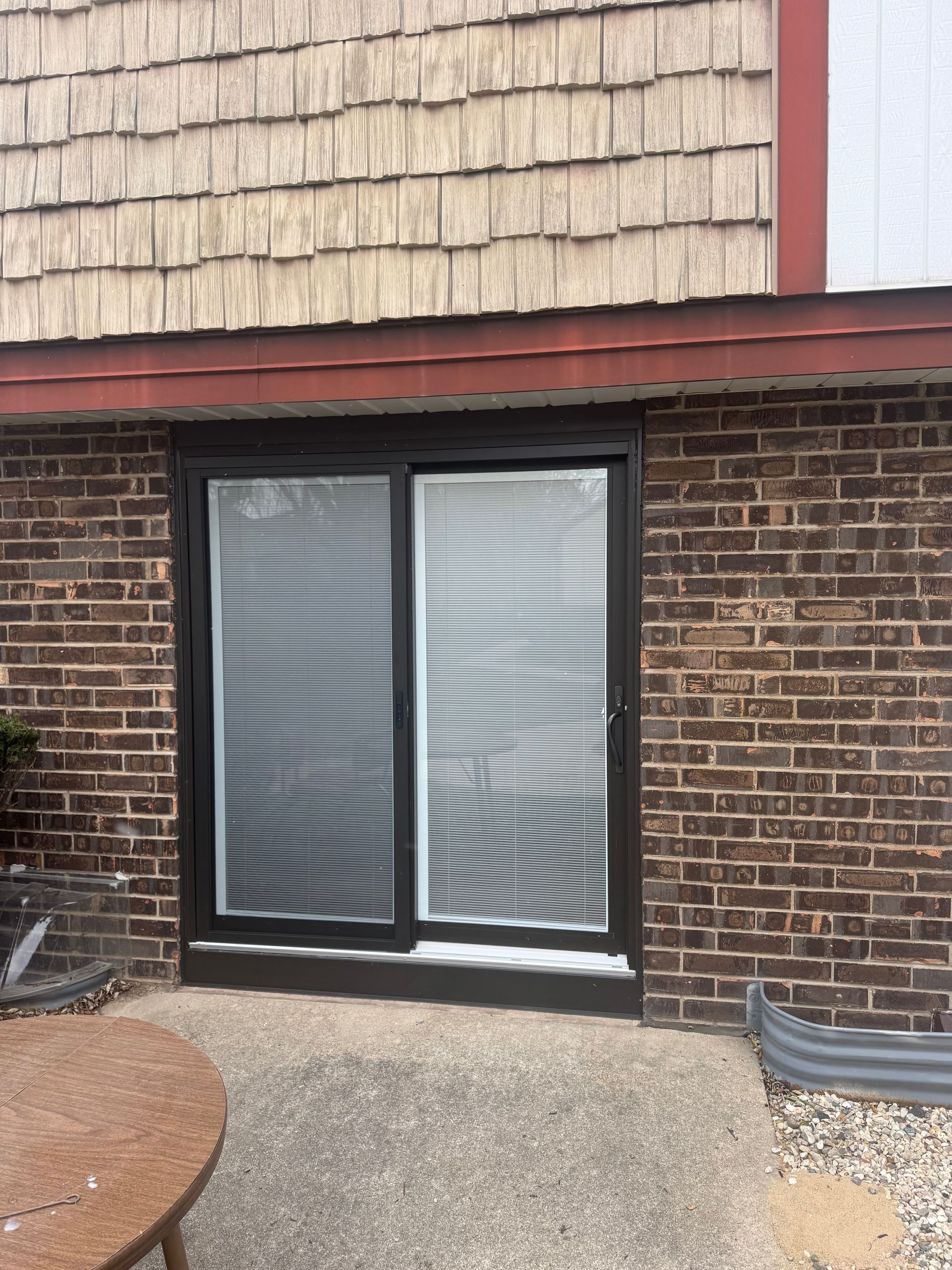 Sliding glass door with blinds, brown brick wall, and textured wood siding. Concrete patio.