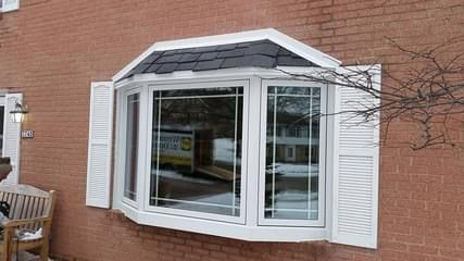 Bay window with white trim, shutters, and slate roof on a brick building.
