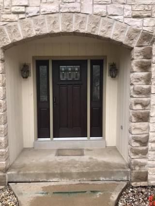 Brown front door with sidelights under a stone archway. Concrete porch with two sconce lights.